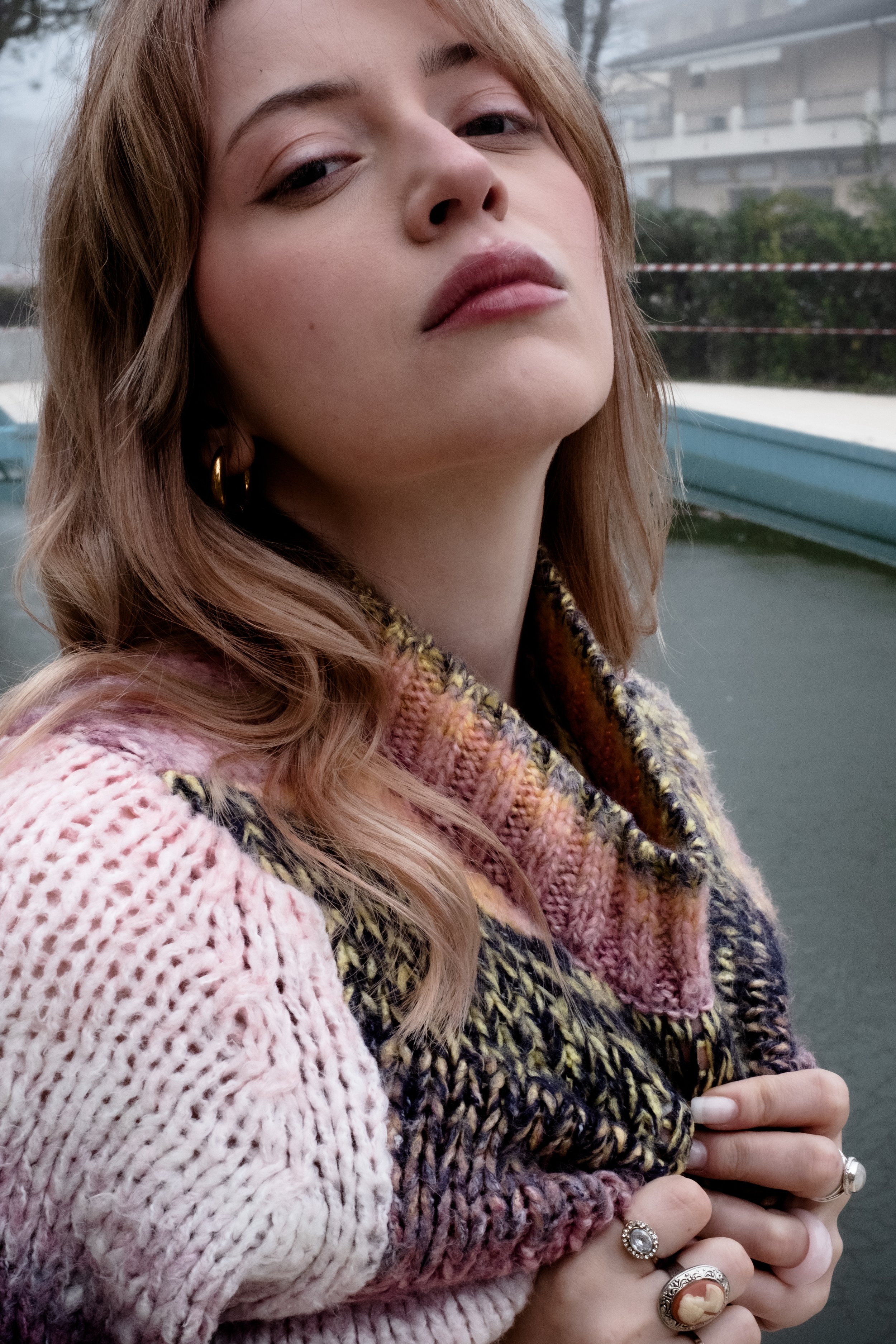 Close-up of a young woman with wavy, shoulder-length hair, wearing a colorful knitted sweater, with her hand showing multiple rings, standing outdoors near a body of water.