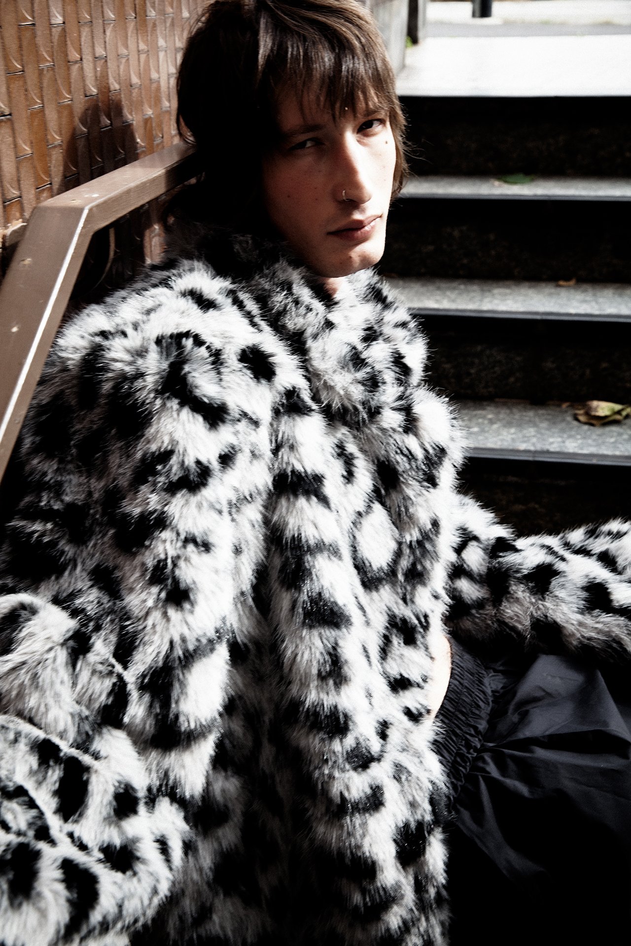 A young person sitting on steps, wearing a black and white spotted faux fur coat, with short brown hair and a nose ring, looking at the camera.