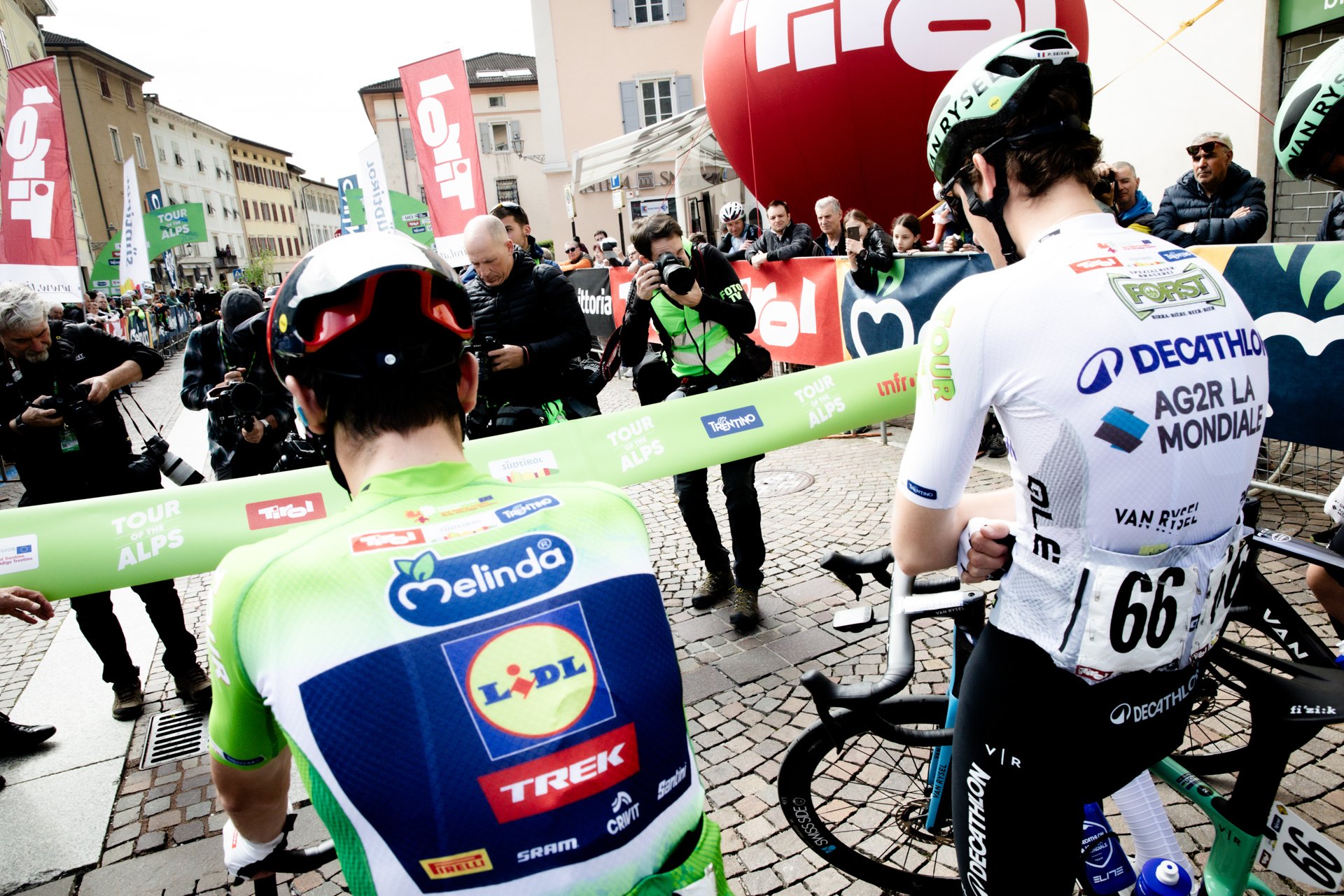 Cyclists at the starting line of the Tour of the Alps with photographers and spectators behind barriers.