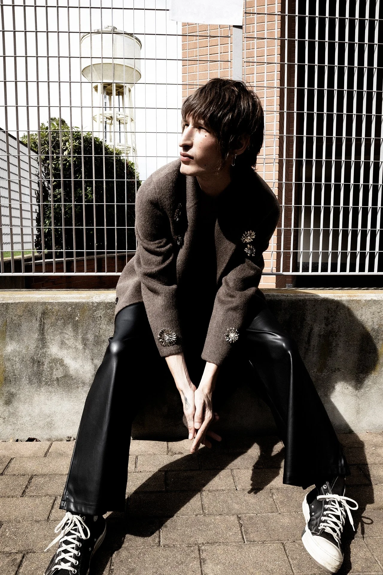 A person squatting outdoors in front of a metal fence and brick wall, wearing a brown blazer with floral embroidery, black leather pants, and black sneakers with white laces, with a water tower visible in the background.