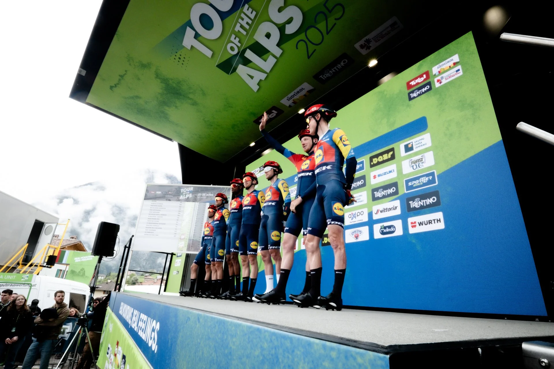 Cyclists in team uniforms standing on a stage, some raising their arms during an award ceremony at the Tour of the Alps cycling race.
