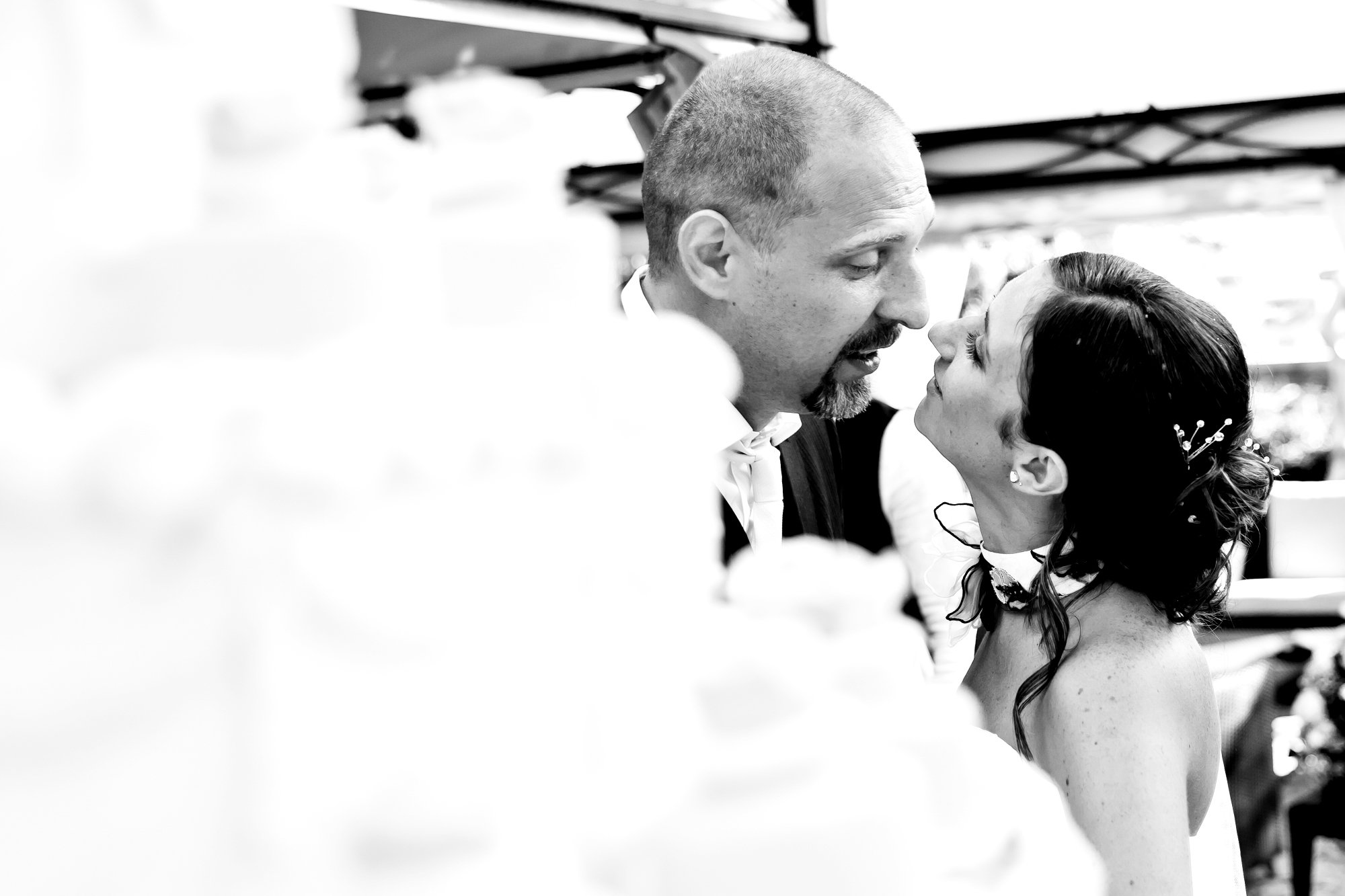 A black and white photo of a man and woman close together, about to kiss, with their noses touching. The woman has dark hair styled with hair accessories and is wearing earrings, while the man has a goatee and short hair. The background is blurred.