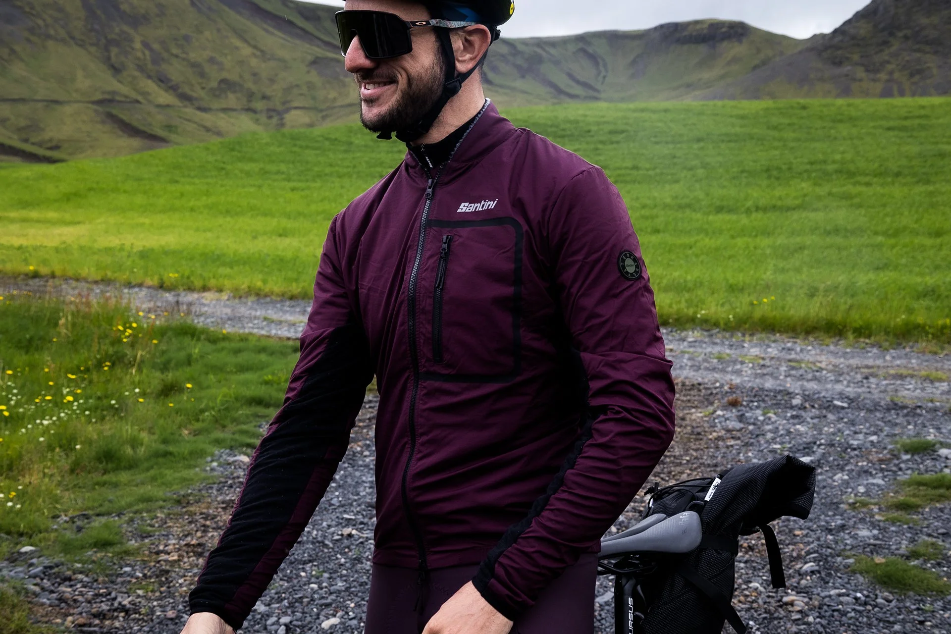 A man in a maroon cycling jacket wearing sunglasses, smiling outdoors on a gravel path surrounded by green grassy hills.