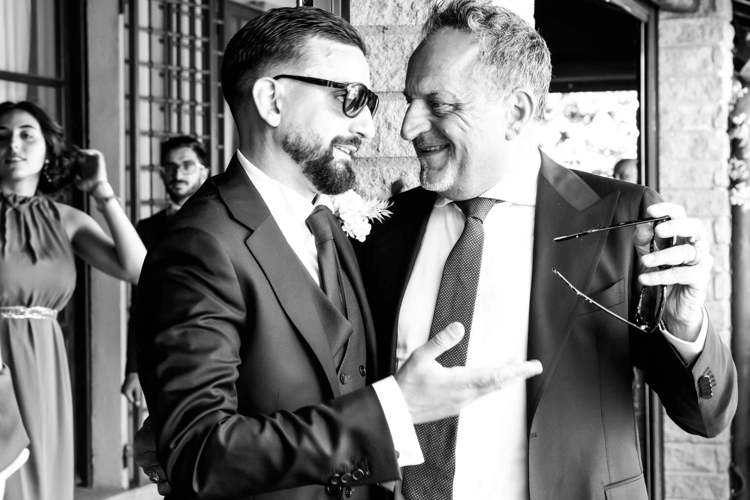 Two men in tuxedos smiling and talking at a wedding reception, with glasses in hand, outdoors near a brick wall.
