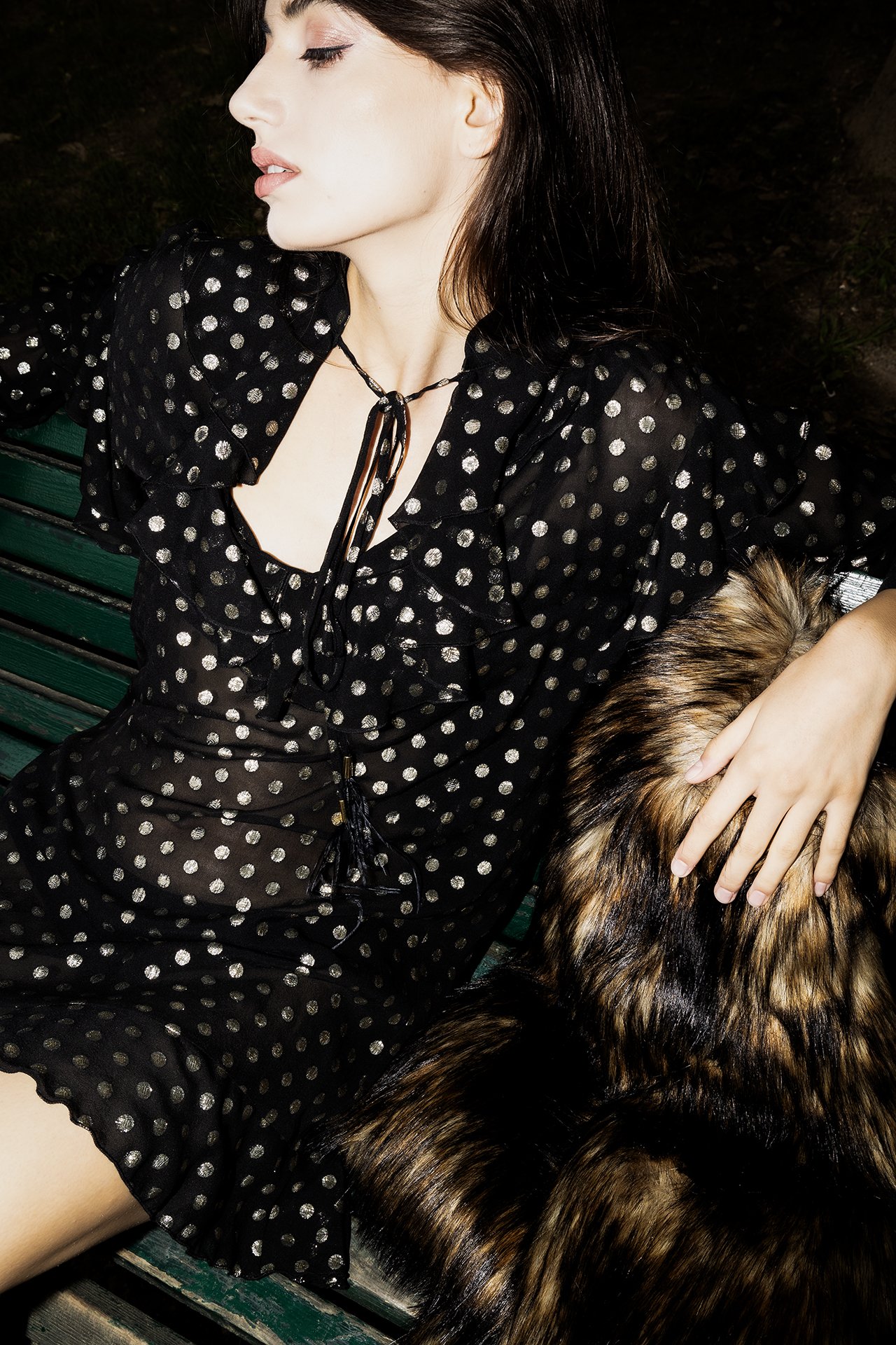 A woman with long dark hair sitting on a green bench, wearing a black dress with silver polka dots, holding a large, multicolored fur coat.