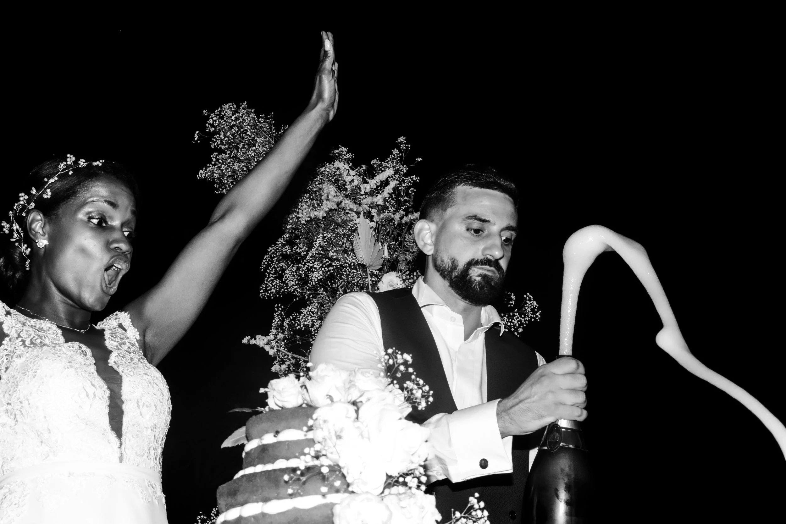 A black and white photo of a bride and groom at their wedding, with the bride on the left raising her arm and the groom on the right using a champagne bottle to pour drink, with flowers and a wedding cake in the background.