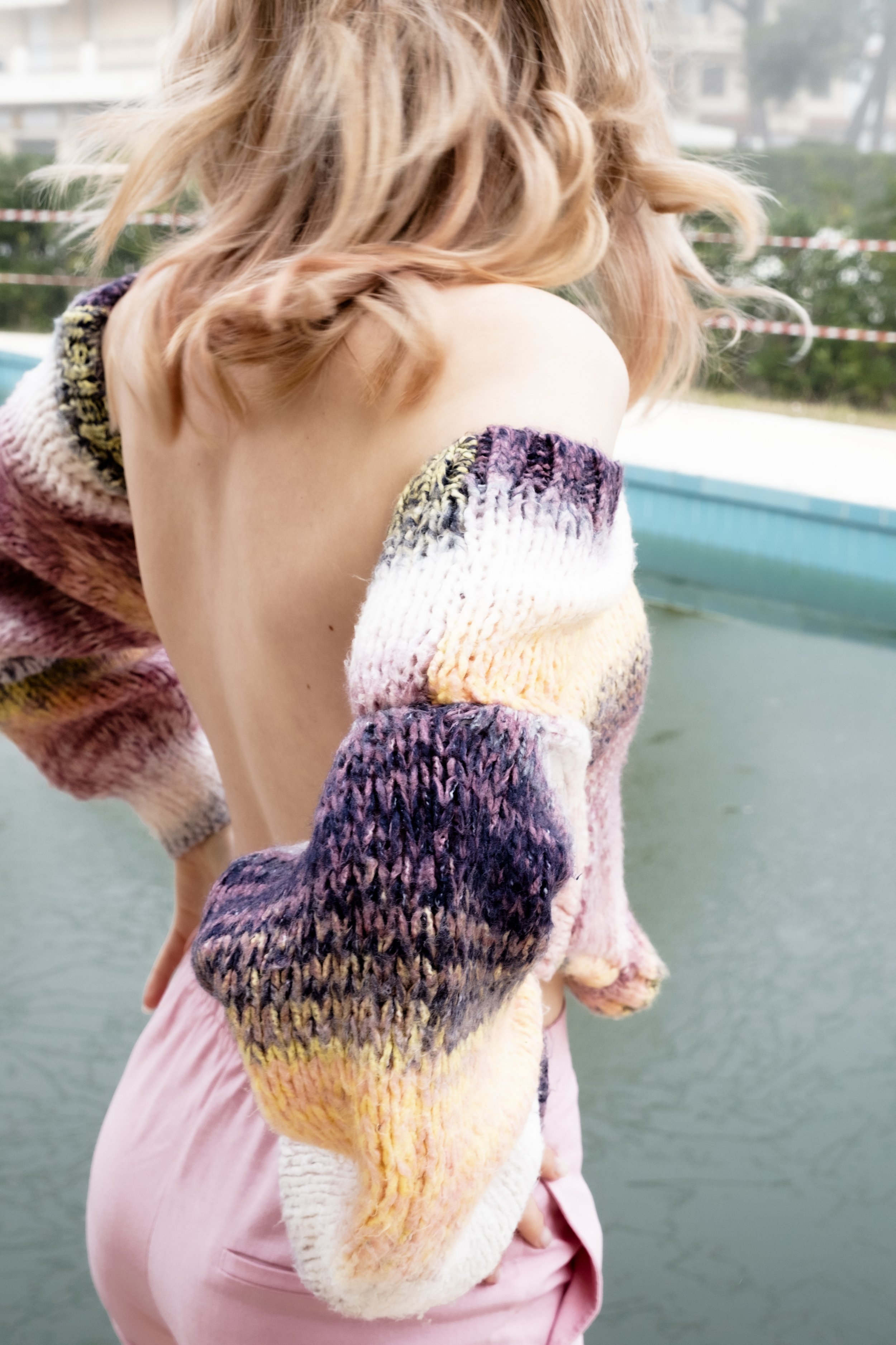 A woman with shoulder-length wavy hair wearing a chunky, multicolored knitted sweater falling off one shoulder, standing near a pool with water in the background.