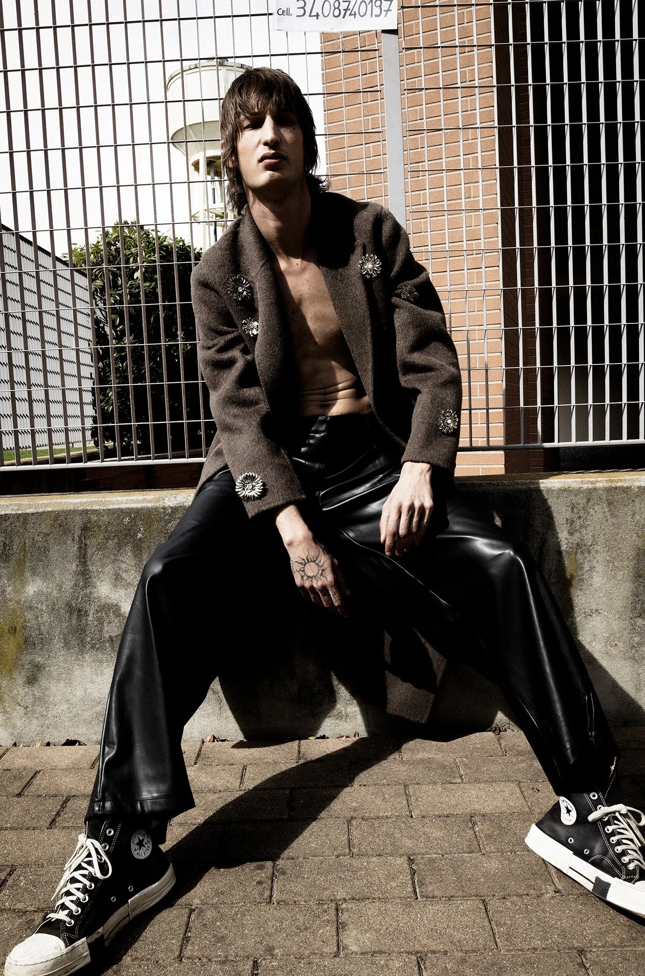 A young adult male with shoulder-length brown hair and tattoos, wearing a decorated brown coat, black leather pants, and black Converse sneakers, sitting on a concrete ledge outdoors near a chain-link fence.