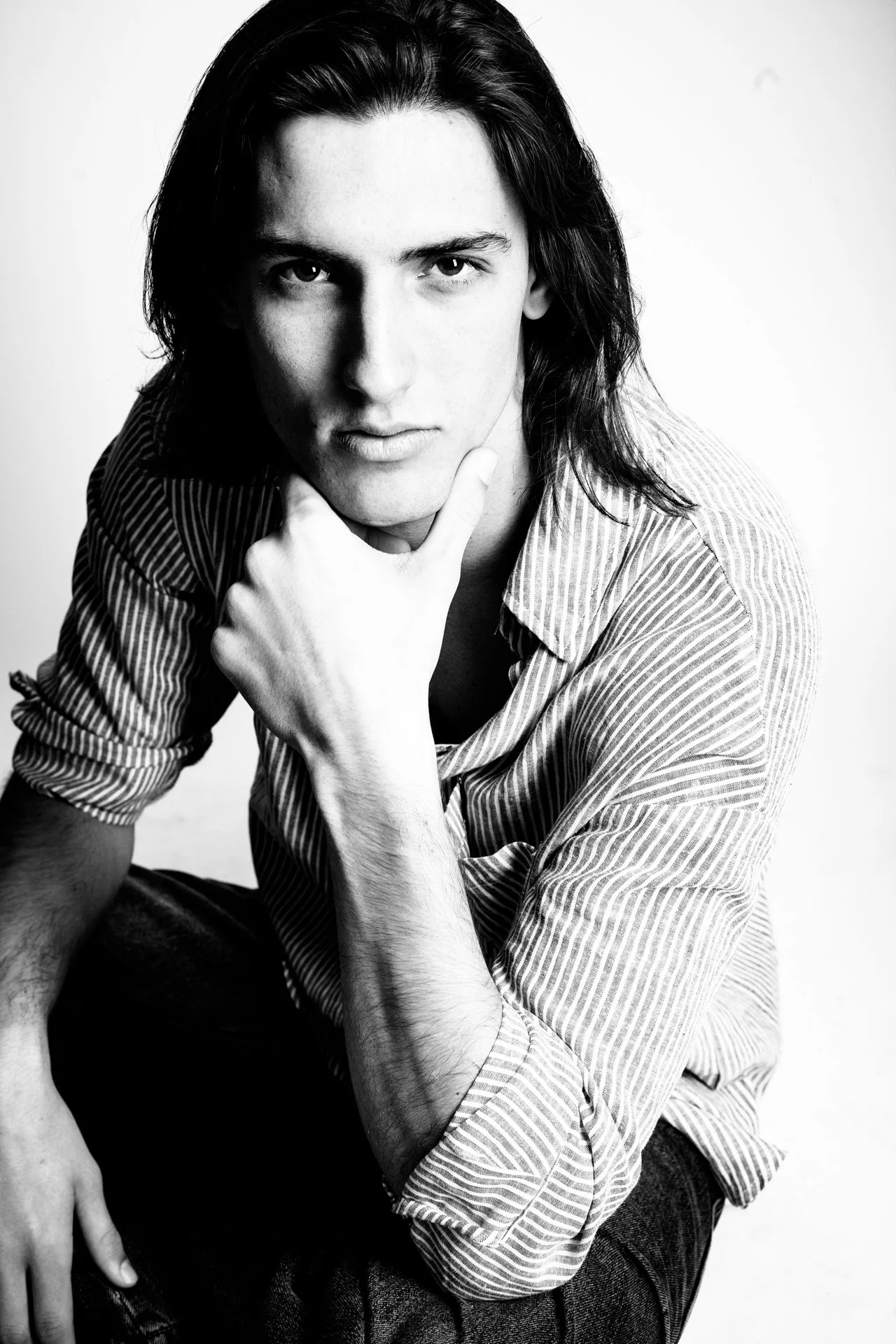 Black and white portrait of a young man with long hair, wearing a striped shirt, looking directly at the camera with one hand touching his chin.