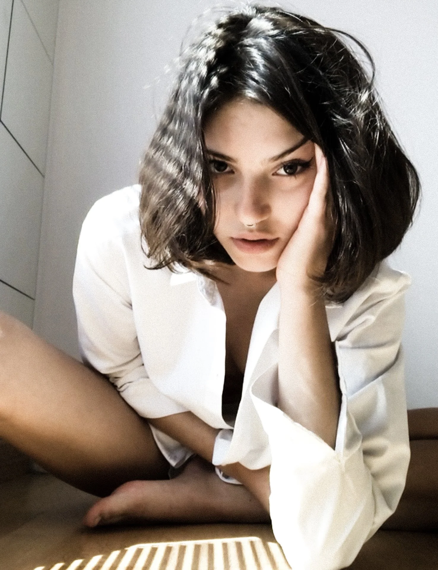 A woman with dark, wavy hair and a nose ring, wearing a white shirt, sitting on the floor with her leg bent, resting her head on her hand, in a room with white walls.