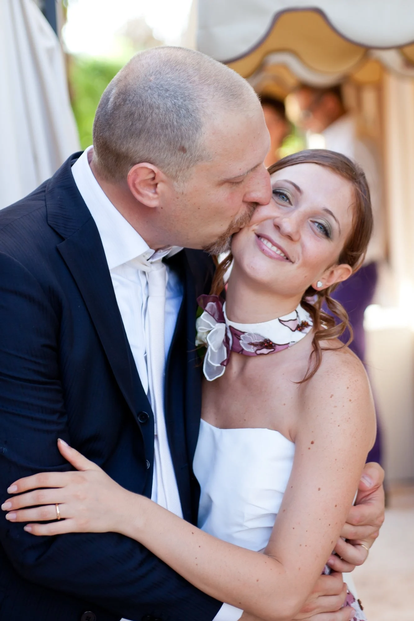A man and woman embrace during a wedding celebration, with the man kissing the woman's cheek while she smiles, dressed in wedding attire, with a decorative arch behind them.