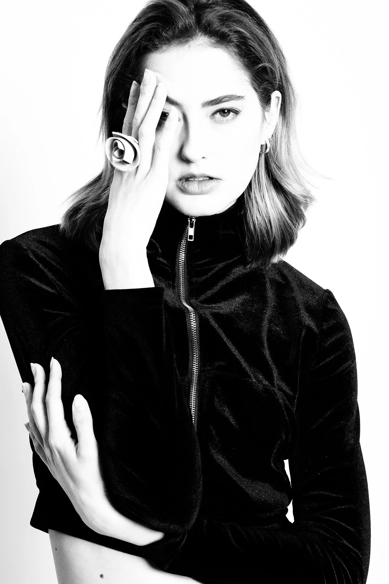 A black-and-white portrait of a woman with shoulder-length hair, wearing a black velvety zip-up top, looking at the camera, with one hand partially covering her face.