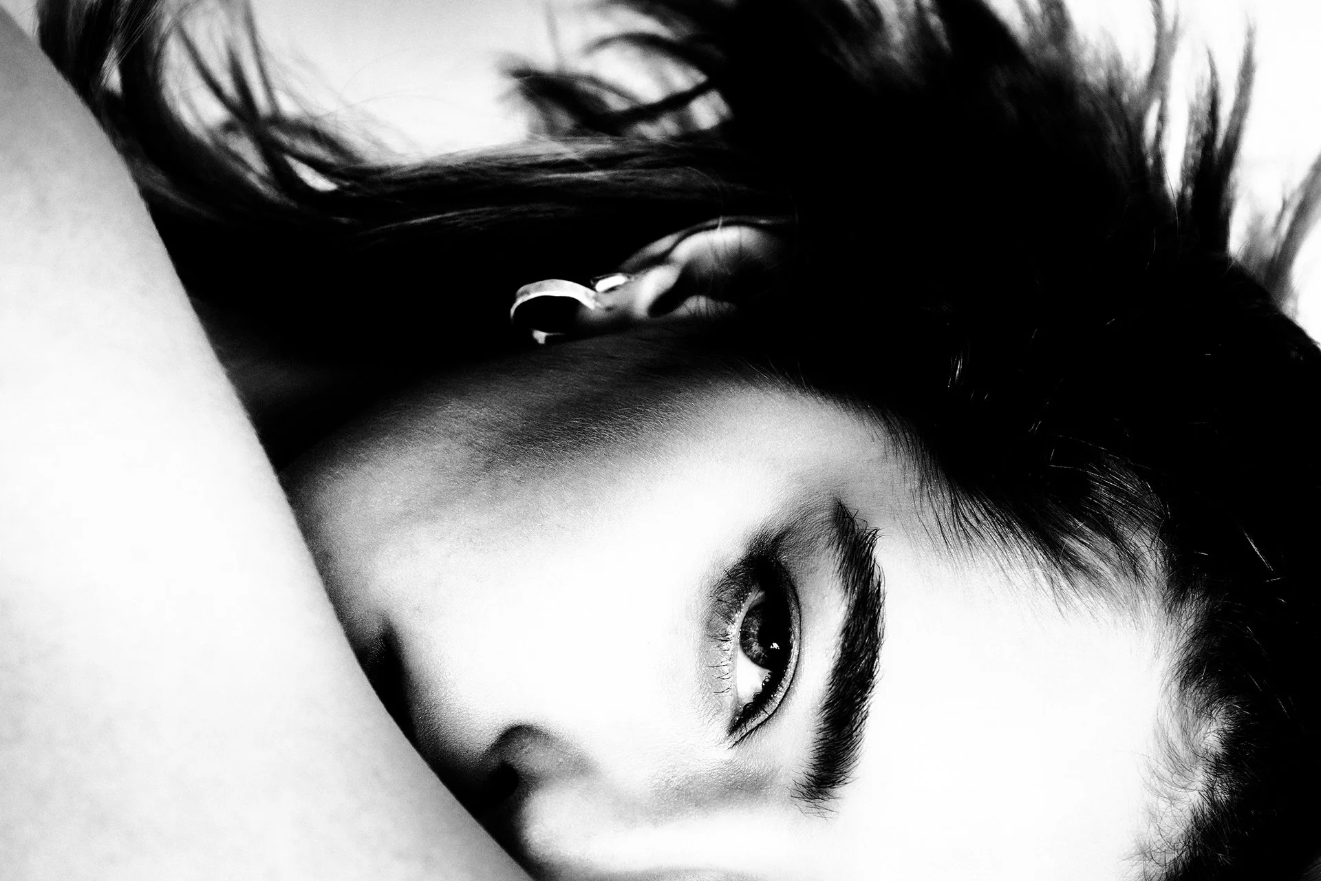 Close-up black and white photograph of a woman's face with dark hair, lying down with only part of her face visible, focusing on her eye and eyebrow.