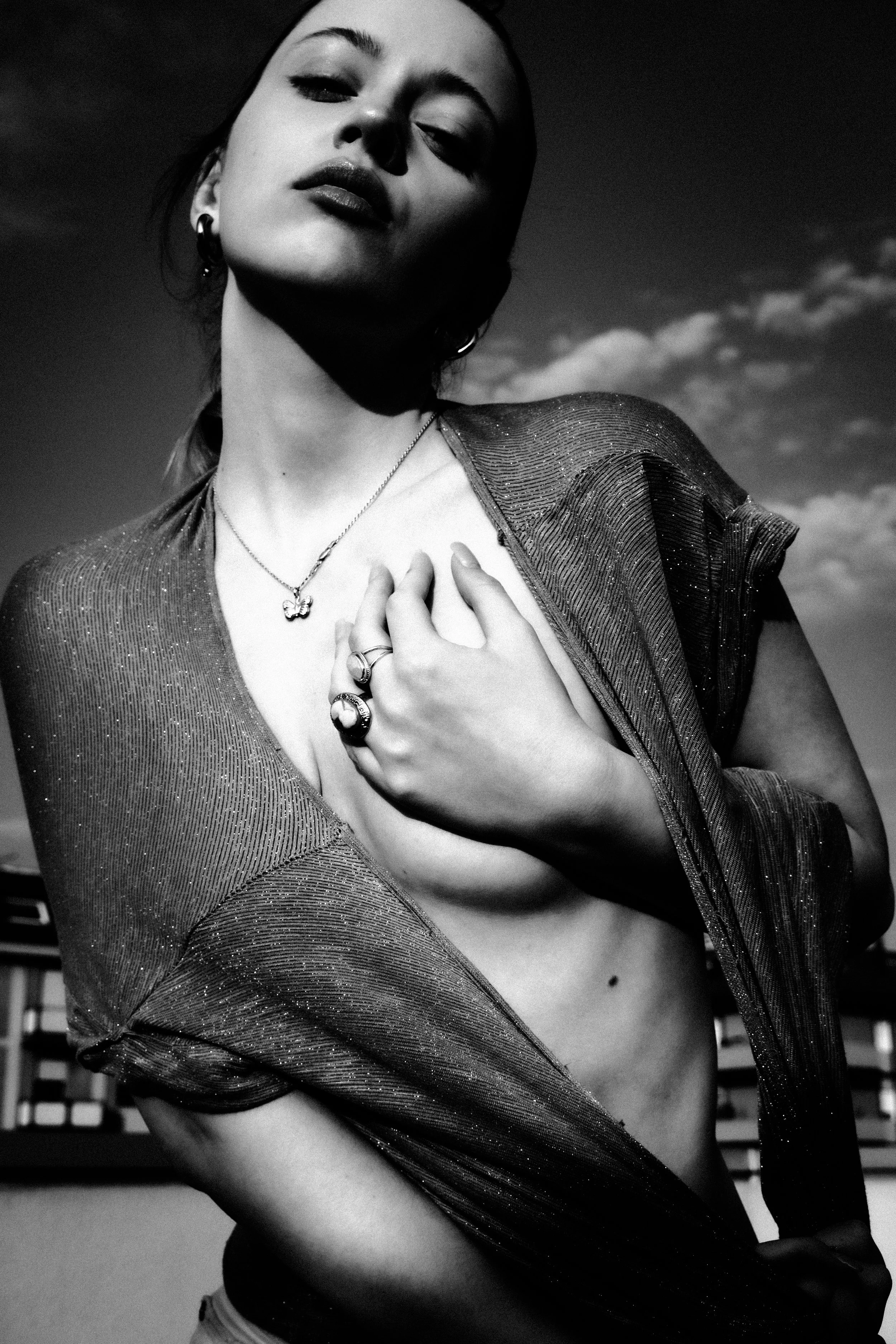 A black-and-white photo of a woman with her head tilted back, wearing a glittery top, jewelry, and holding her chest with one hand while exposing part of her midriff. She has a confident expression, and a cloudy sky is visible in the background.