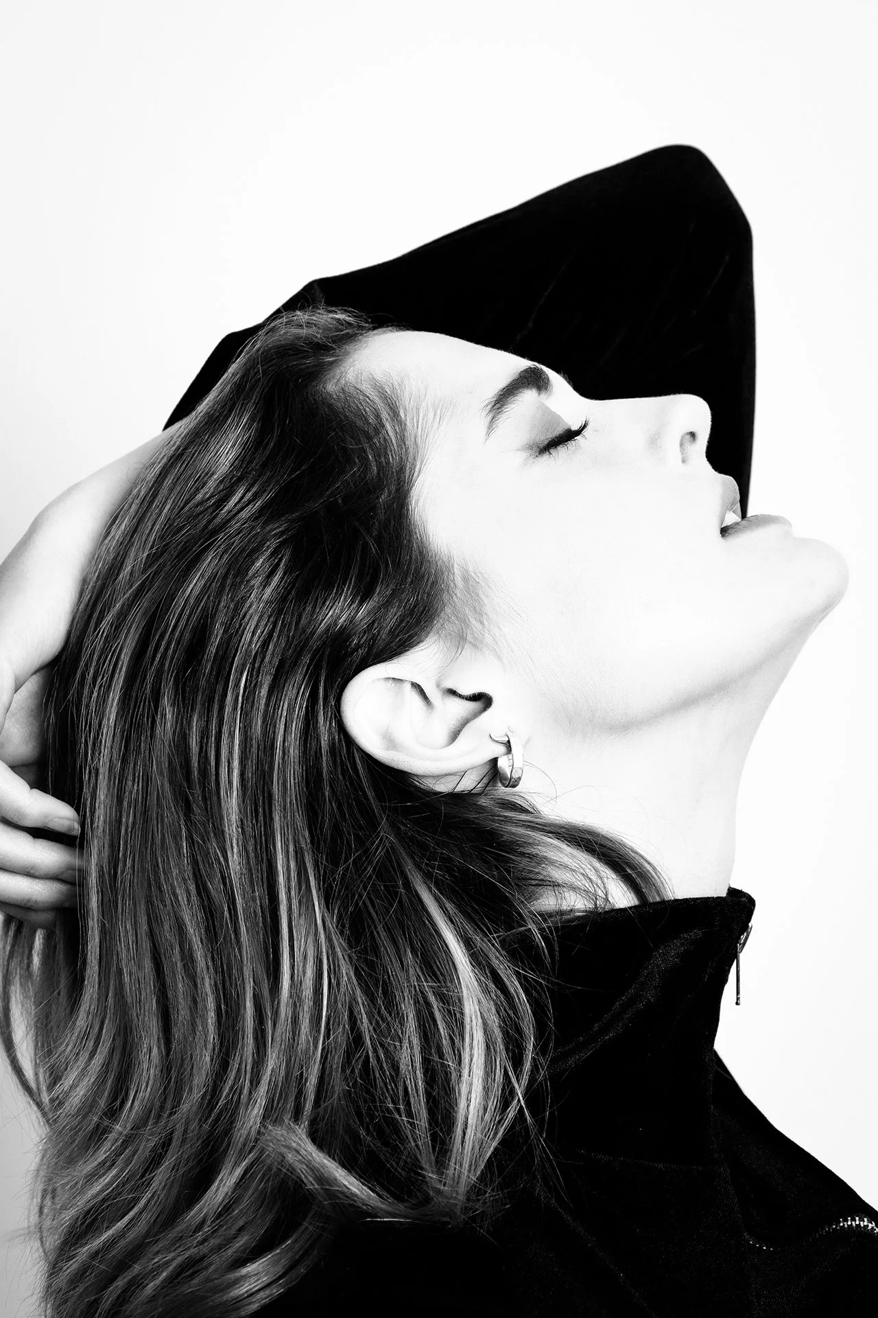 A black and white portrait of a woman with closed eyes, head tilted back, and hand resting on her head. She is wearing a jacket with a zipper and hoop earrings.