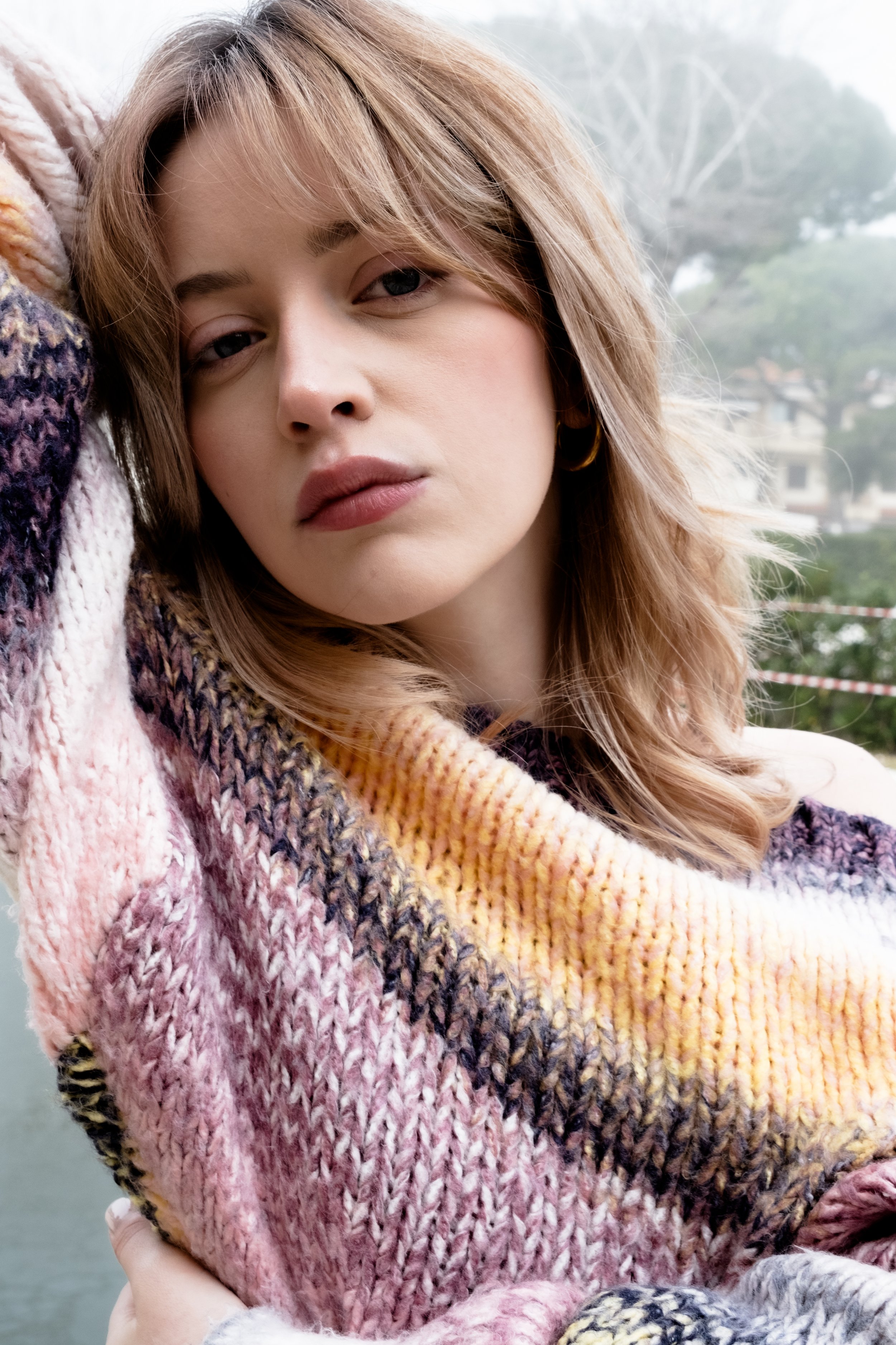 A young woman with blond hair and hoop earrings posing outdoors, wrapped in a multicolored knit blanket.