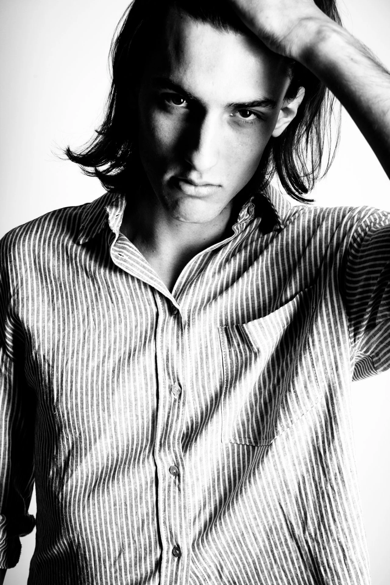 A black and white portrait of a young man with shoulder-length hair, wearing a vertically striped shirt, looking directly at the camera with one hand touching his head.
