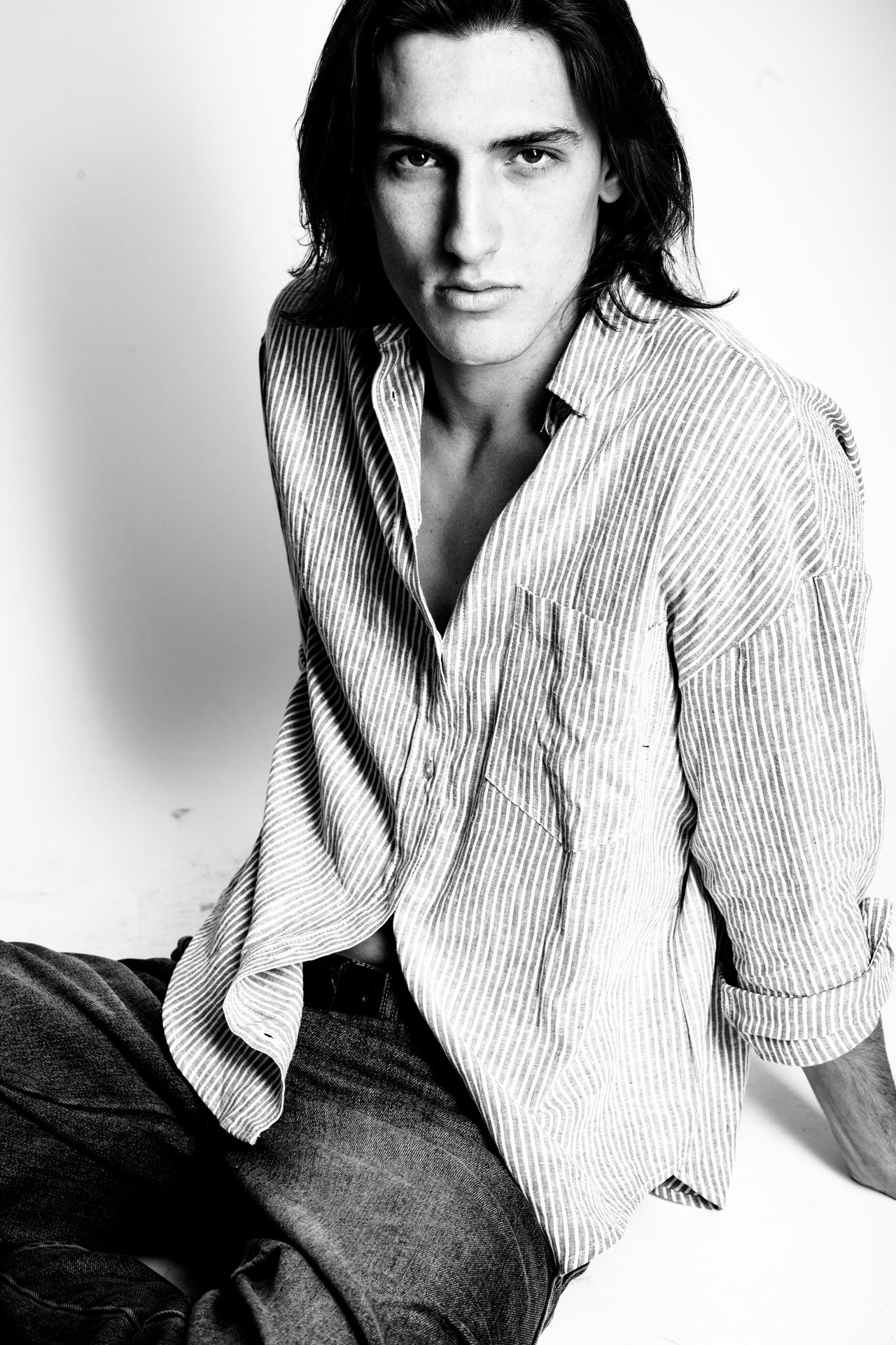 Black and white portrait of a young man with long dark hair, wearing a striped shirt, sitting against a plain background.