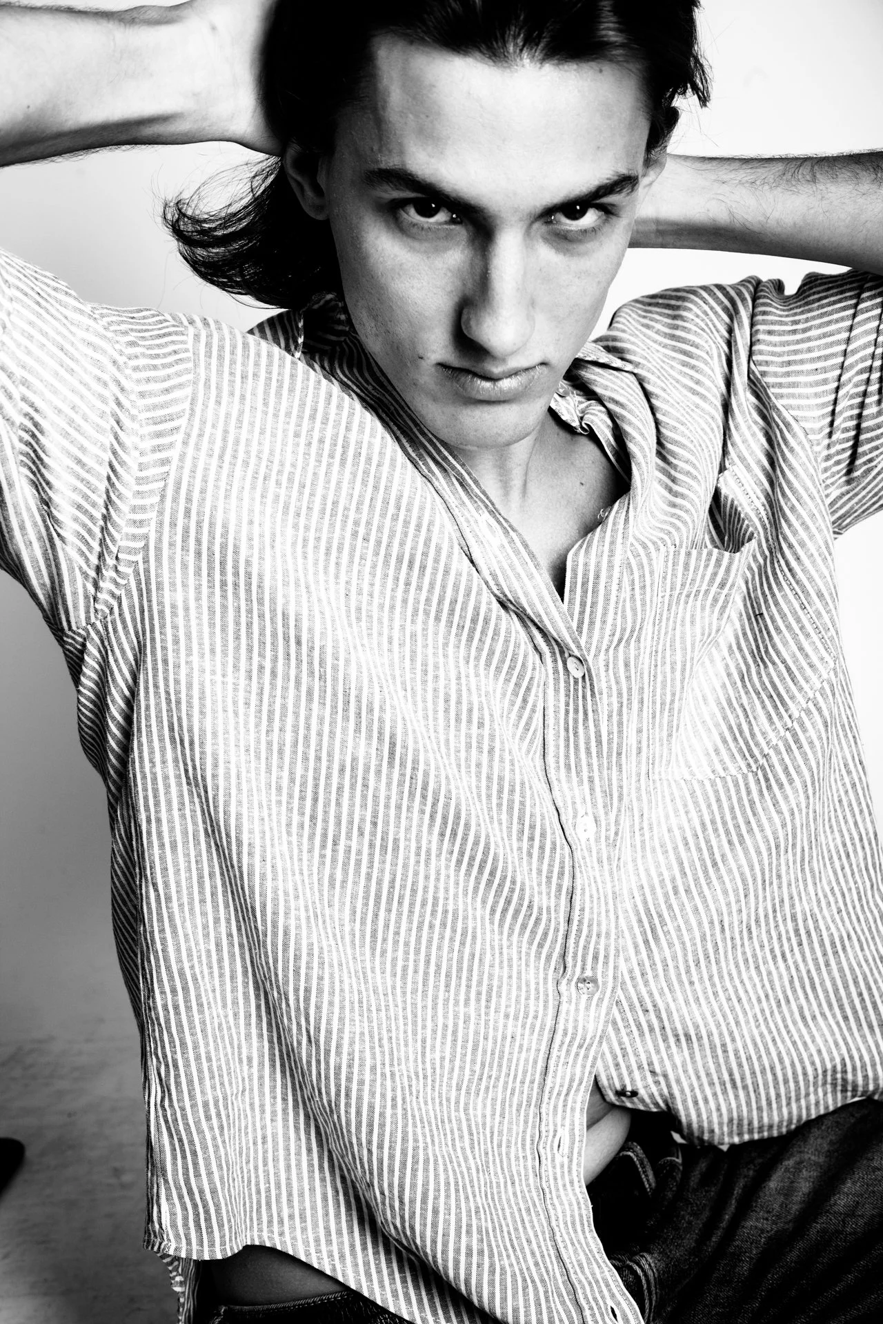 Black and white photo of a young man with dark hair, wearing a striped button-up shirt, looking intensely at the camera with hands behind his head.