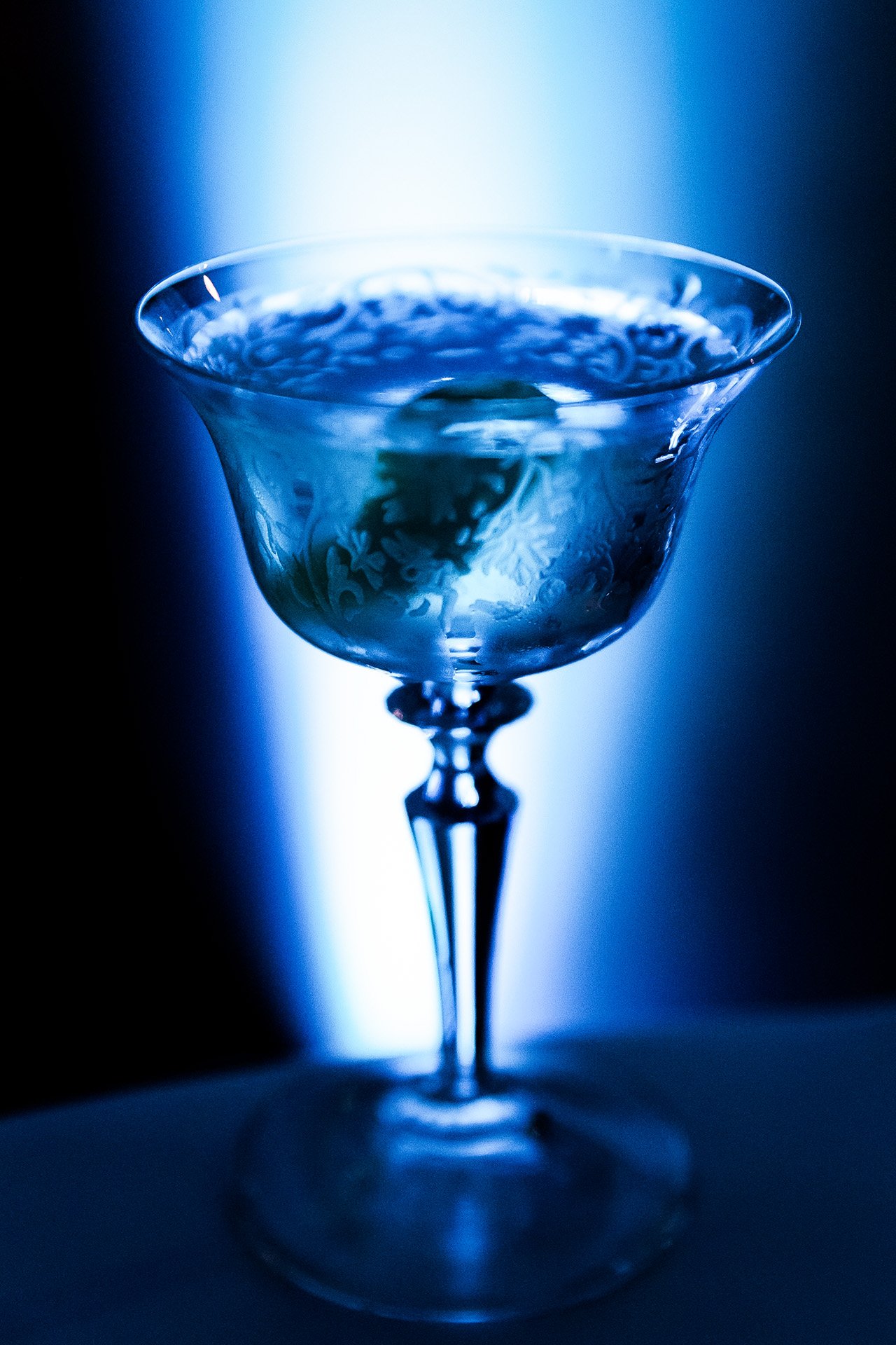 A close-up of a clear cocktail glass with water and a lime wedge inside, illuminated by blue light in a dark setting.