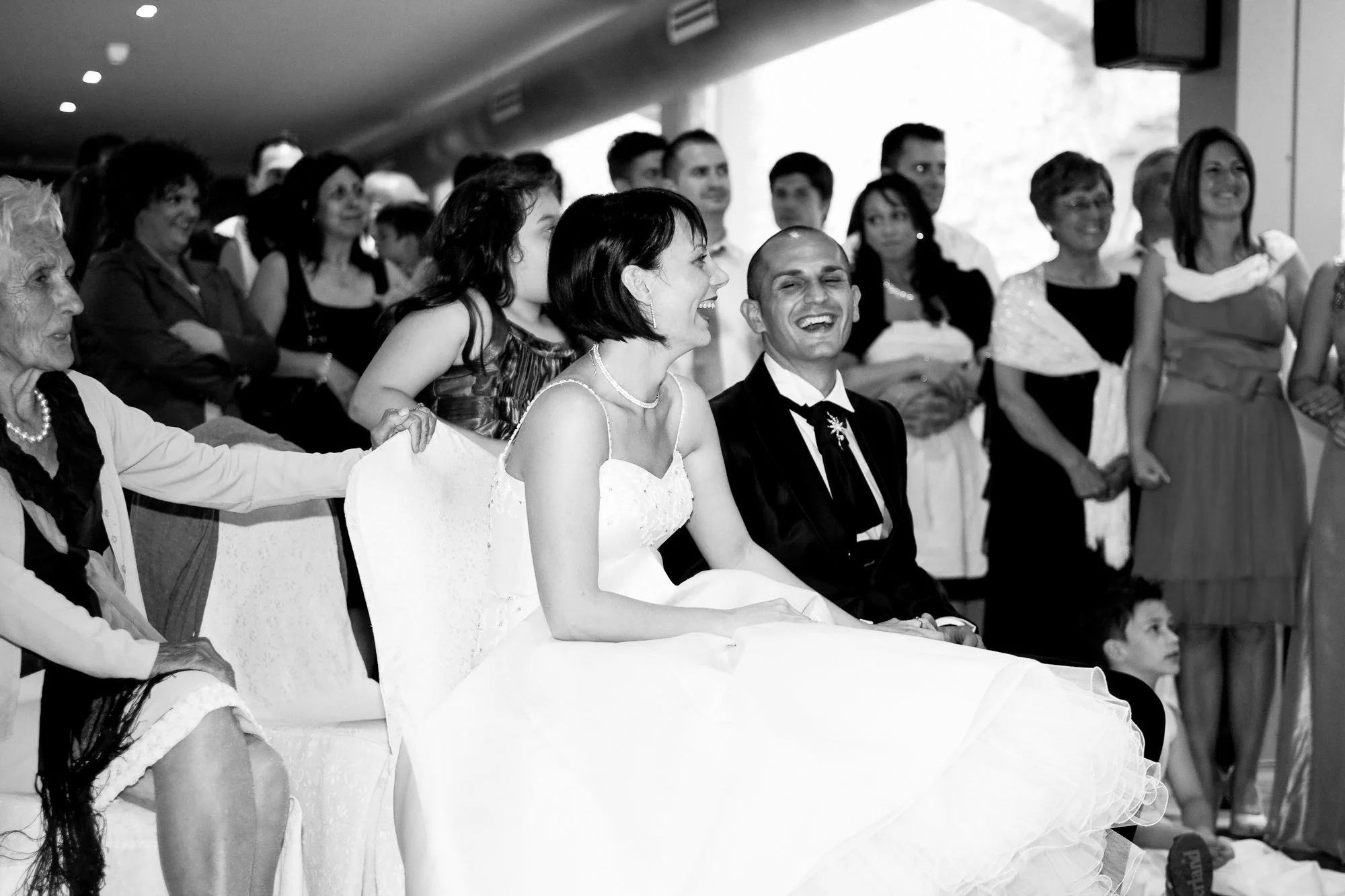 A bride and groom sitting together, surrounded by a group of people during a wedding celebration.