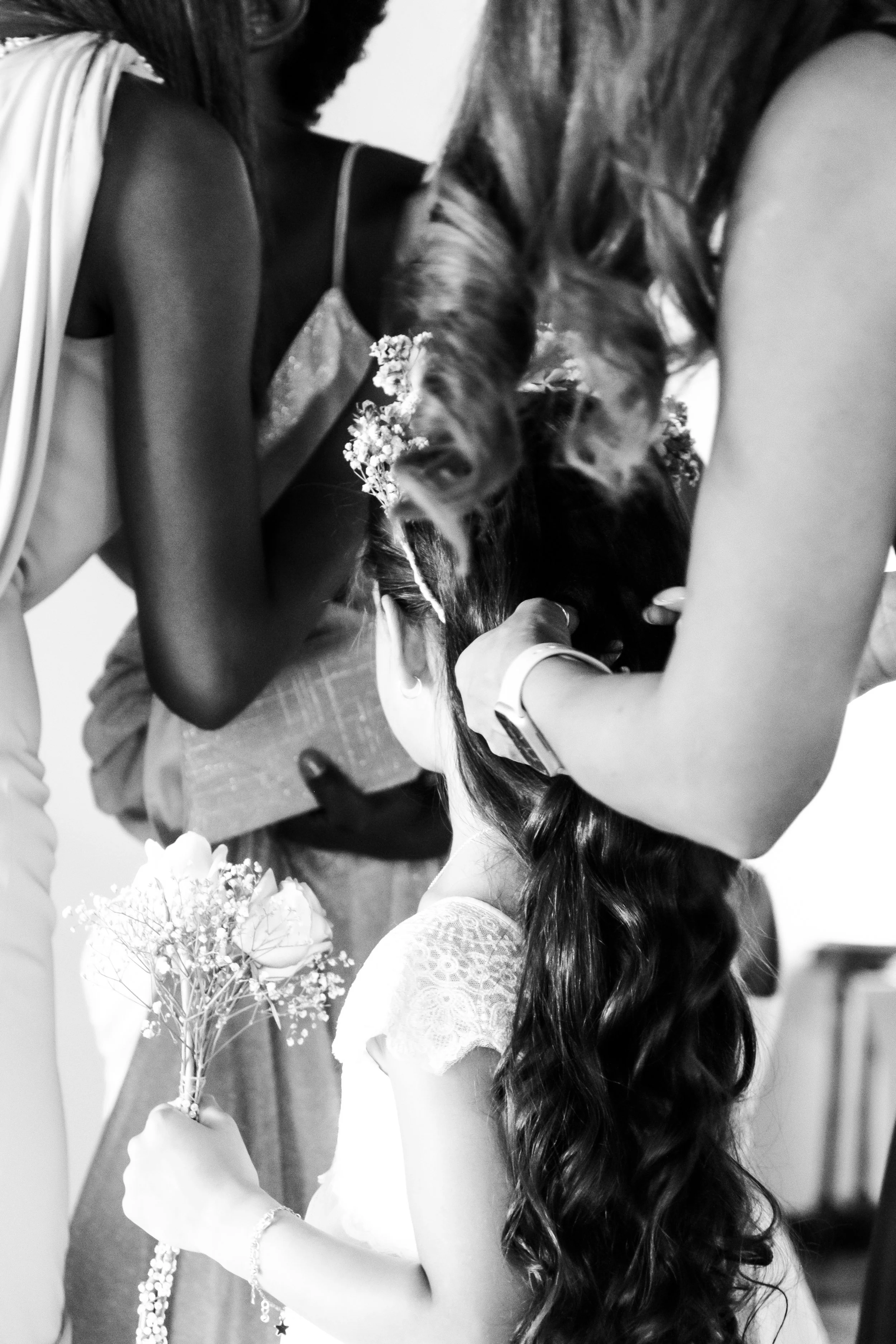 A girl with long curly hair and lace dress holds a small bouquet of flowers while two women lean over her, adjusting her hair and head, during a wedding or similar event.