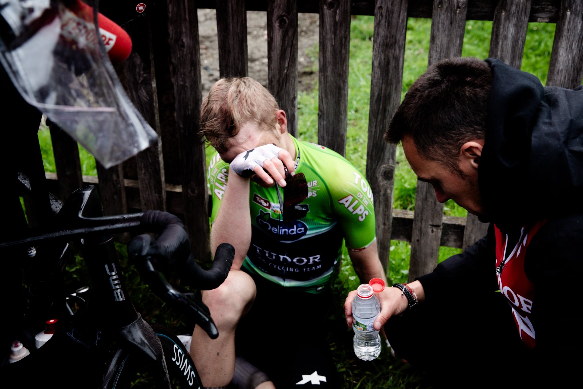 A cyclist in a green jersey is sitting on the ground, wiping his face with a towel, appearing exhausted. A team member is offering him a water bottle, and a bicycle is visible nearby.