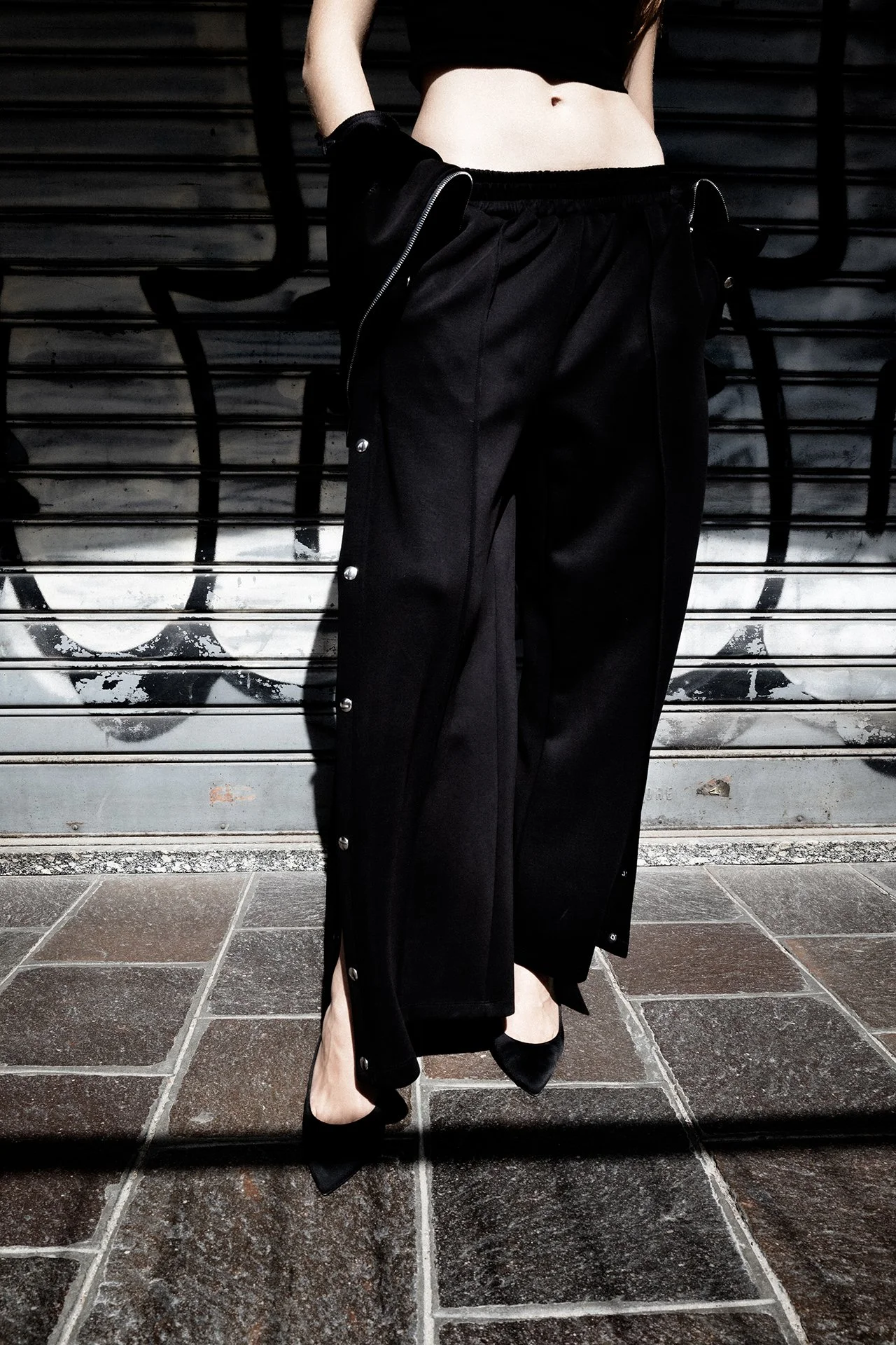 A person wearing black pants, high heels, and a black top, standing against a graffiti wall with graffiti and horizontal lines.