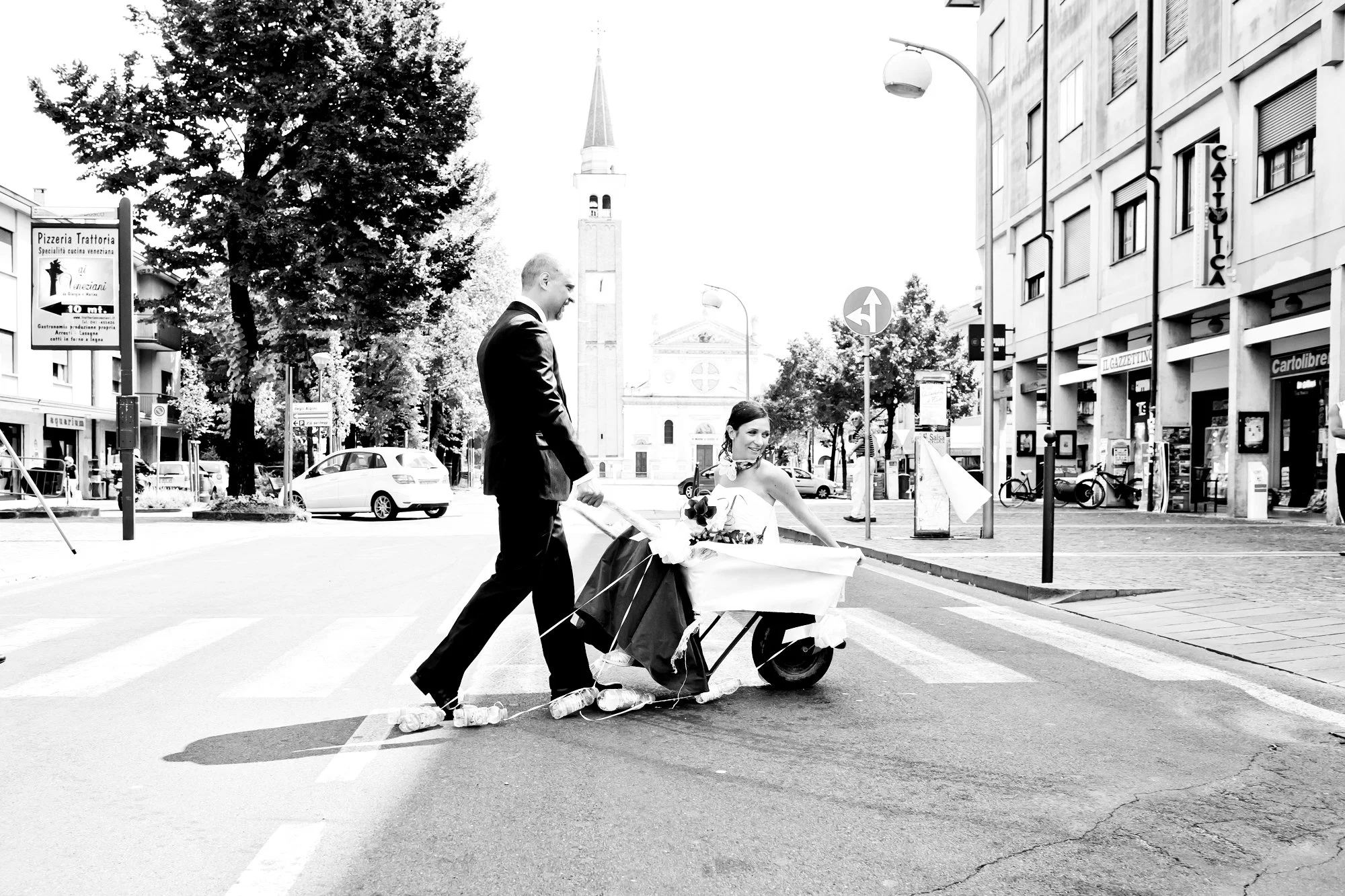 A black and white photo of a man in a suit pushing a woman in a wedding dress on a scooter across a city street. The woman holds a bouquet of flowers and is smiling. Buildings and cars are visible in the background.