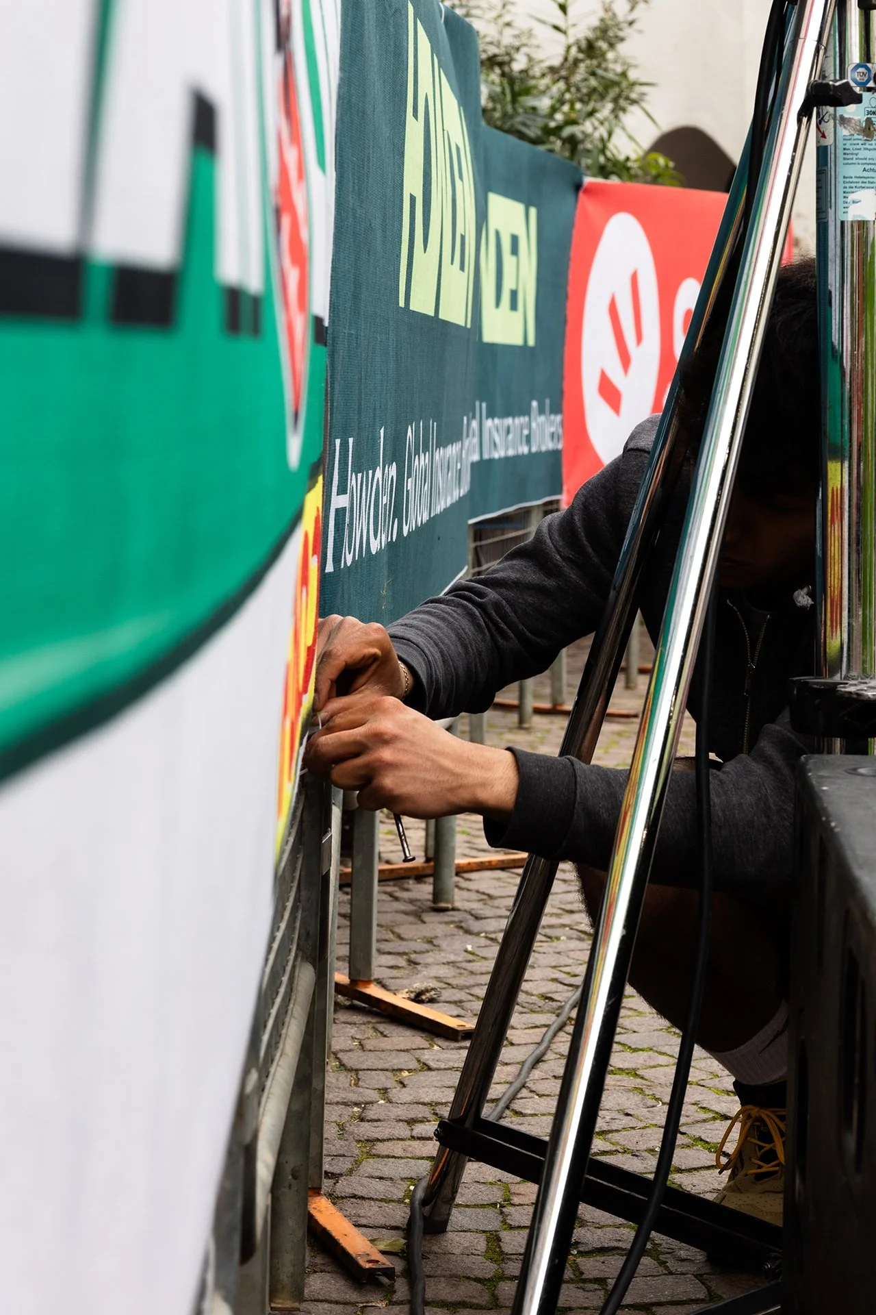 A person is attaching a sticker to a large banner with a tool, during an outdoor event on cobblestone ground.