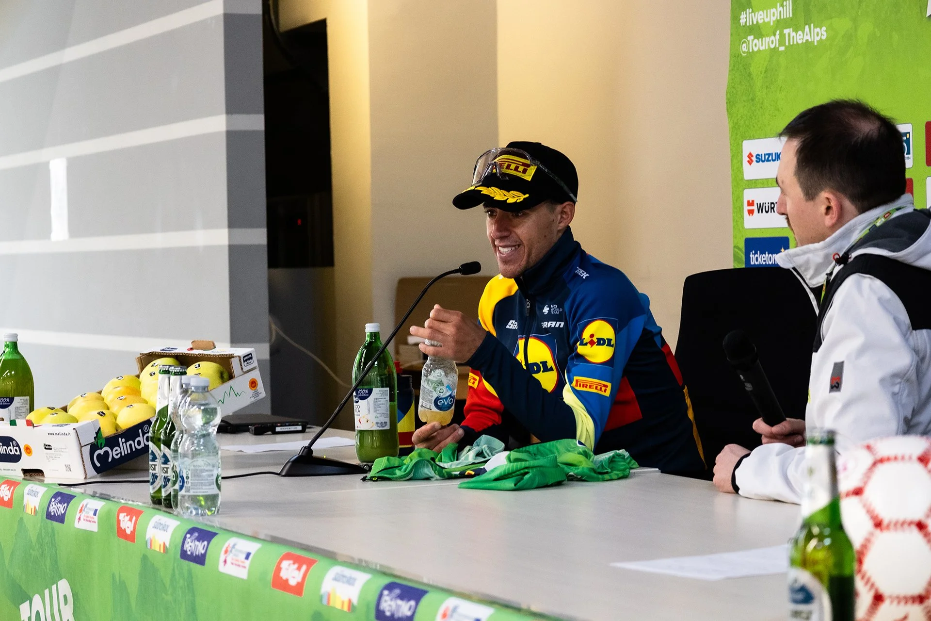 A man in a racing suit and cap sitting at a press conference table, smiling and holding a water bottle, with another man holding a microphone sitting nearby.