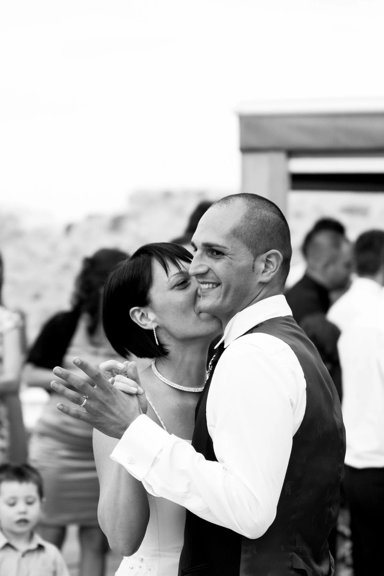 A black-and-white photo of a bride and groom dancing closely and smiling during their wedding reception. The bride has short dark hair and is wearing earrings and a pearl necklace. The groom has a shaved head and is dressed in a white shirt with a da