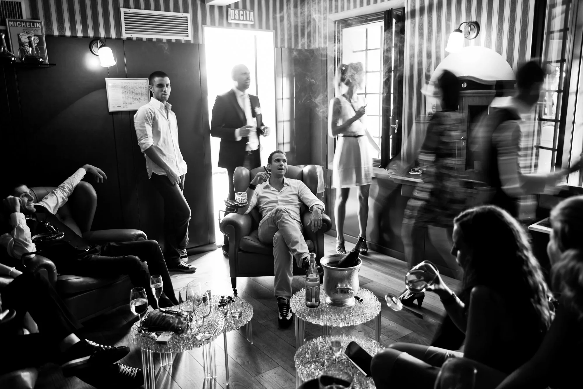 A black and white photo of a lively social gathering in a cozy indoor setting, featuring people seated and standing, with some holding drinks and engaging in conversation.