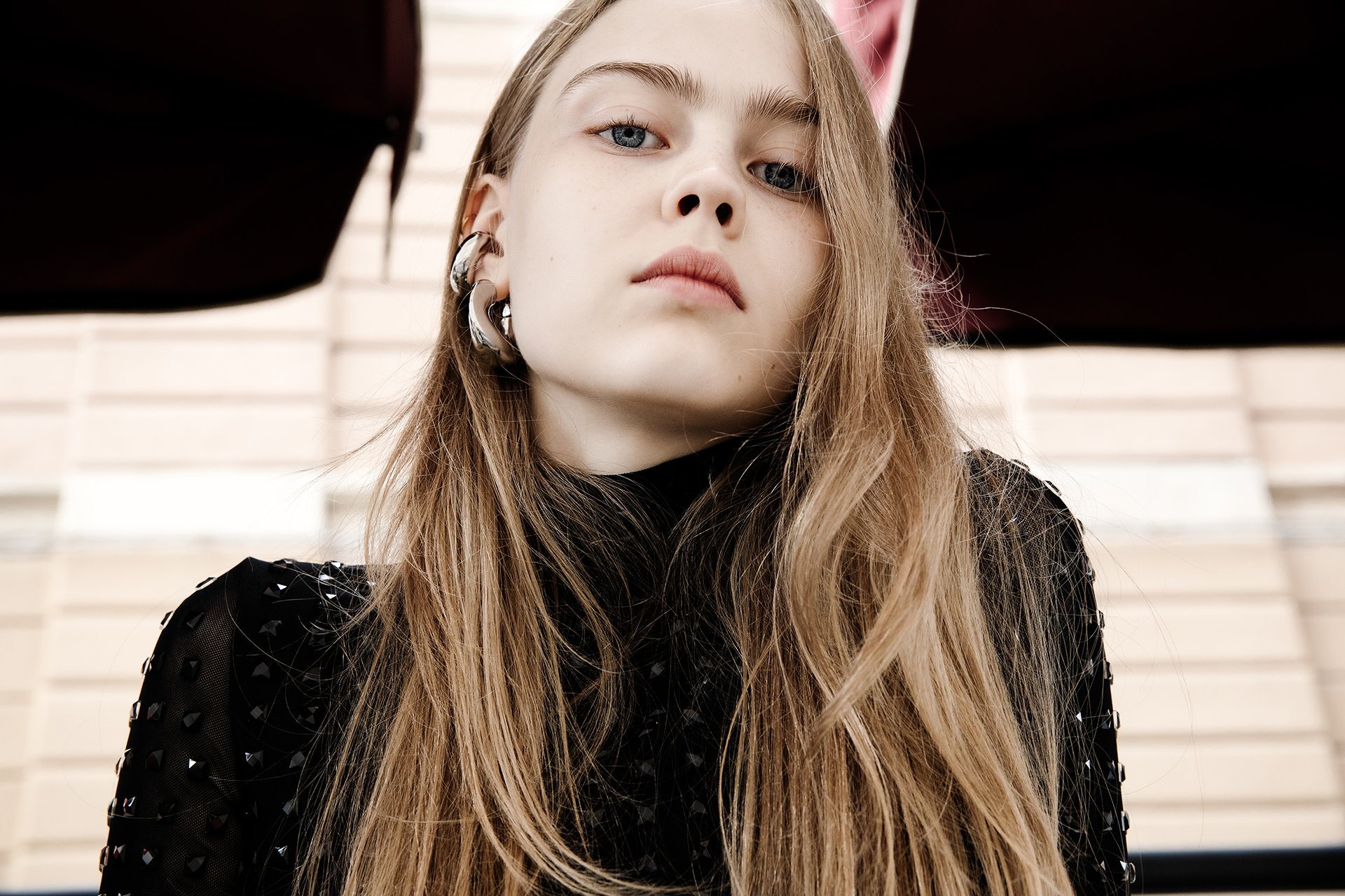 Close-up of a young woman with long, wavy blonde hair and blue eyes, wearing multiple large earrings and a black top with decorative details, looking at the camera with a serious expression.
