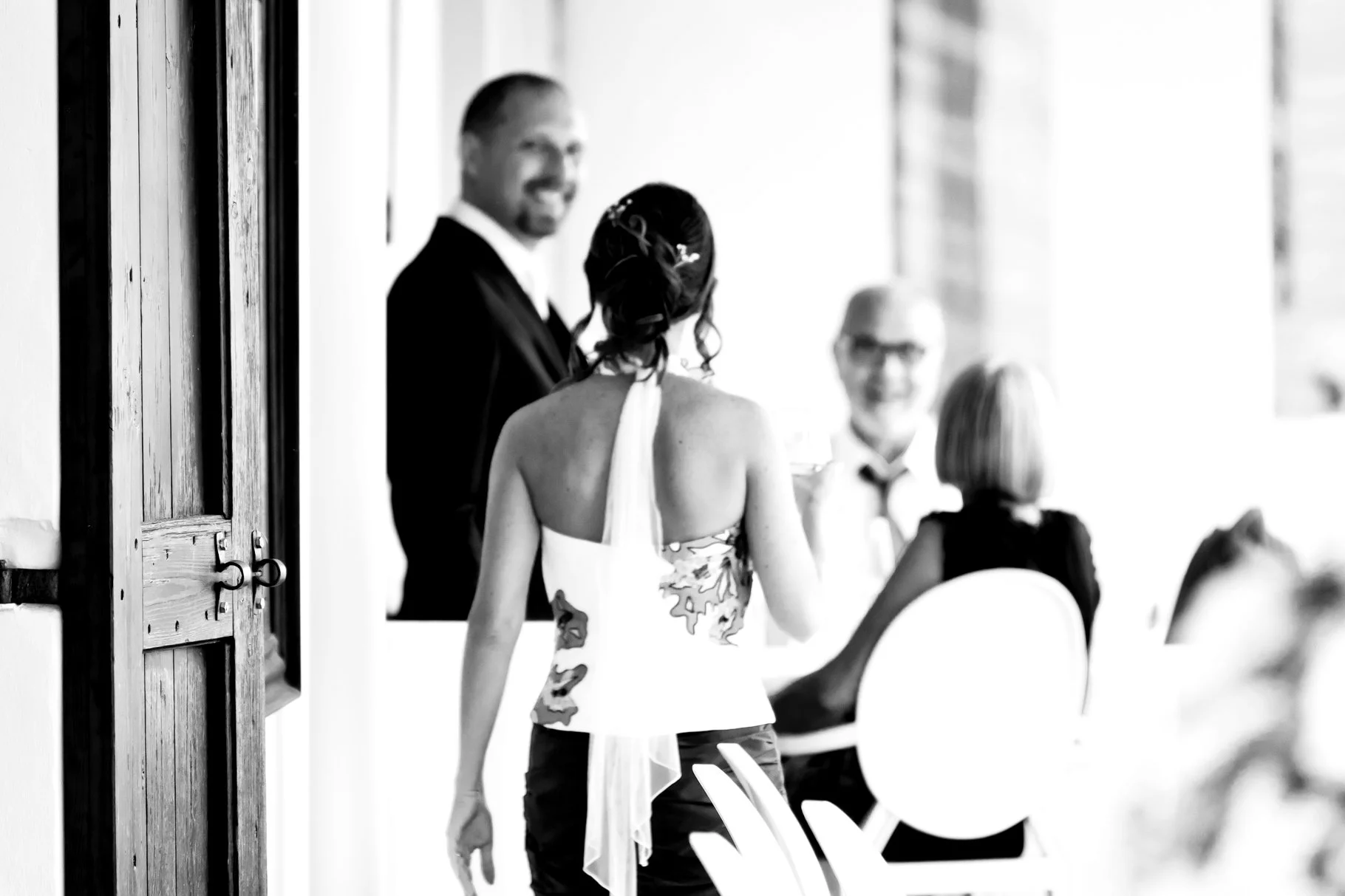 A woman in a sleeveless dress stands with her back facing the camera, talking to a man in a tuxedo at a formal event, with two other people sitting at the table. The scene is in black and white.