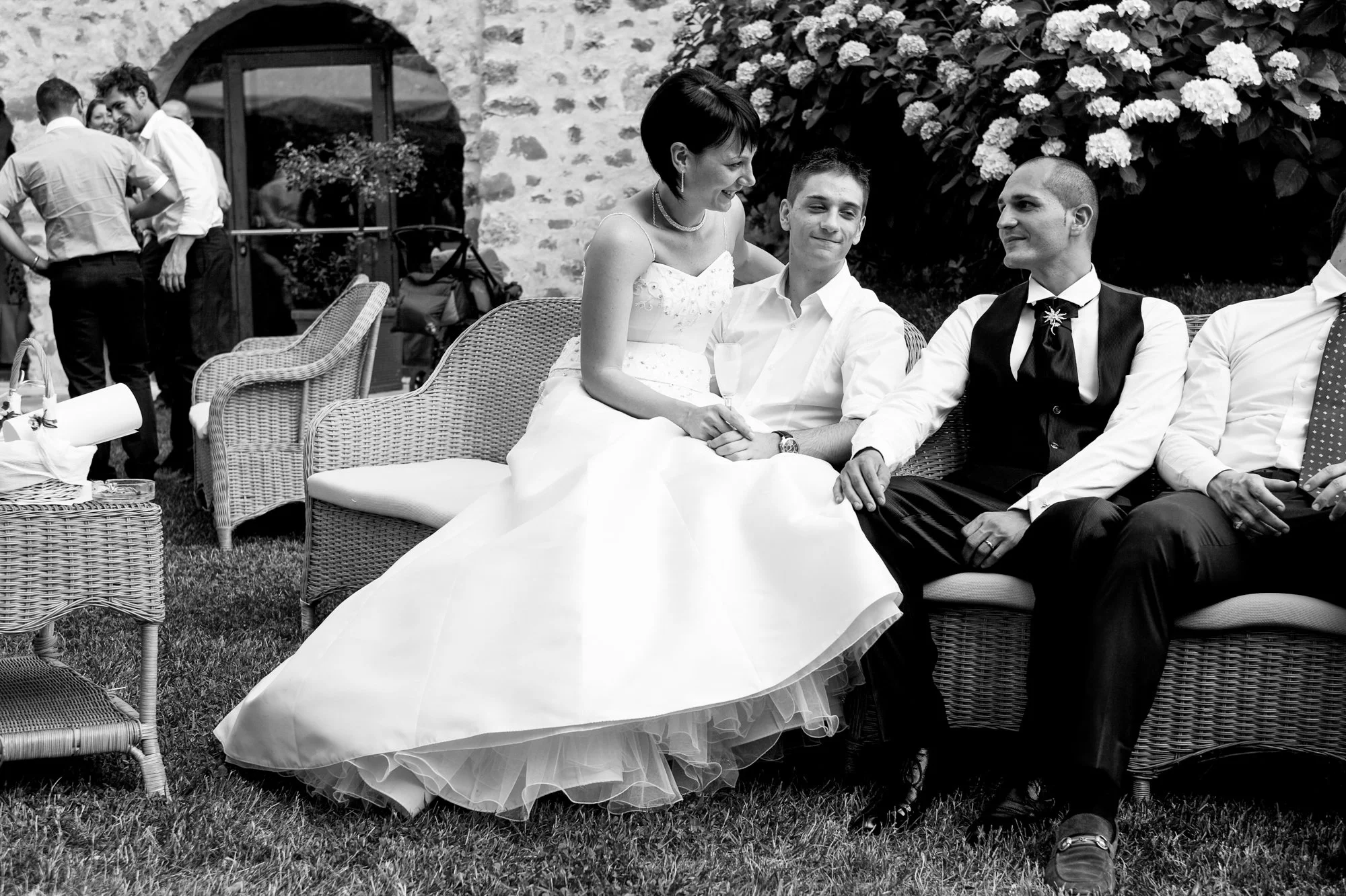 Black and white photo of a wedding celebration with a bride sitting on a wicker sofa, smiling and talking with two men sitting beside her, all dressed in formal attire. In the background, several people are standing and socializing near an arched sto