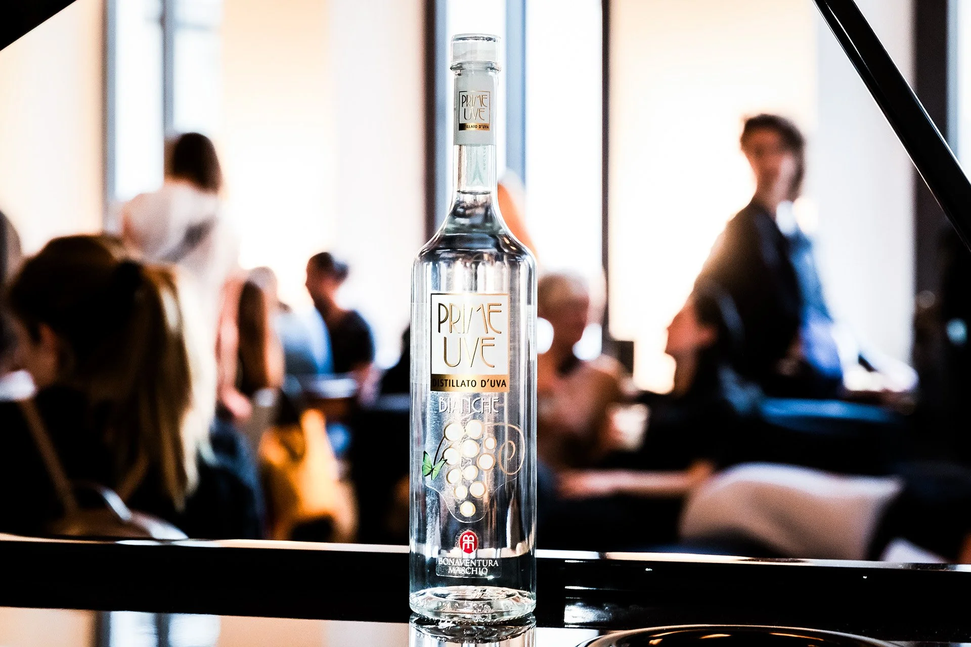 A clear glass bottle of Privé Uve grape distillate on a black surface, with a blurred crowd of people in a well-lit room in the background.