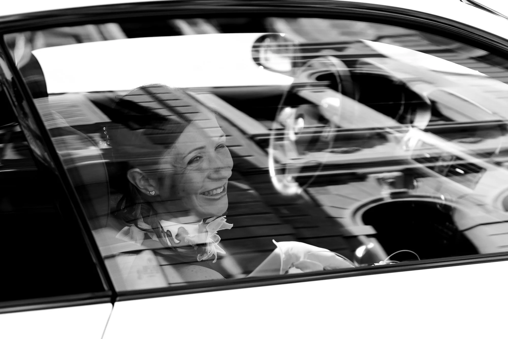 A woman sitting in the driver's seat of a car, smiling and looking out the window, with reflections of buildings and the sky on the car's rear window.