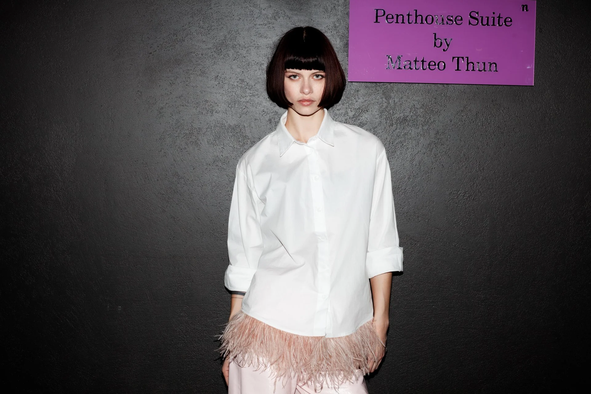 A woman with dark brown bob haircut and bangs wearing a loose white shirt with rolled-up sleeves, standing against a black textured wall. To her right, there is a purple sign with black text that reads 'Penthouse Suite by Matteo Thun.'