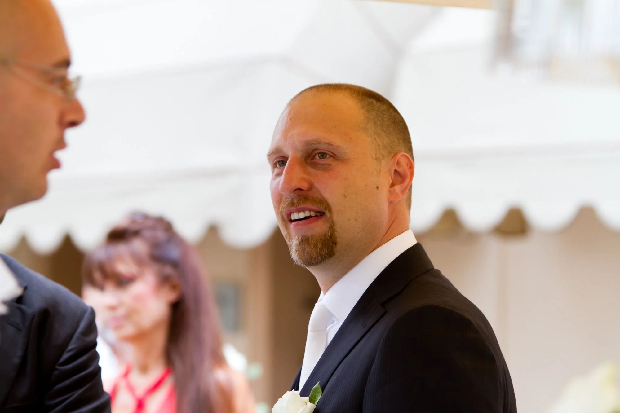 A man in a black tuxedo with a white shirt and tie, with a boutonniere, is looking at another person during a wedding or formal event.