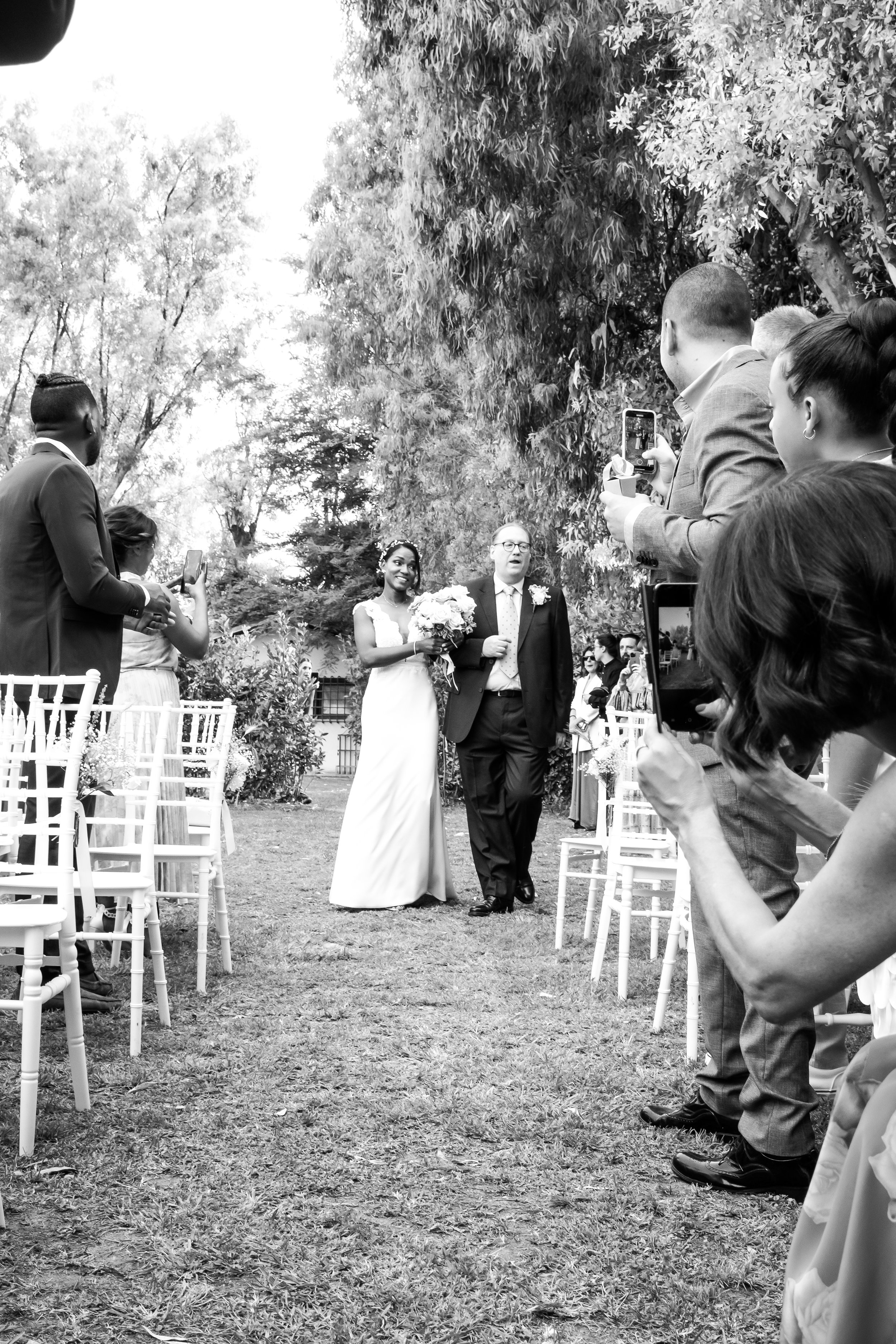 Black and white photo of a wedding ceremony outdoors with the bride and a man walking down the aisle, surrounded by guests taking photos.
