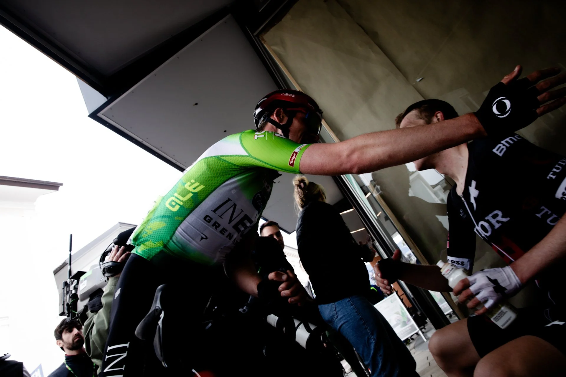 A cyclist in green and white kit talking to a team member in black kit inside a team area, with other staff and riders in the background.
