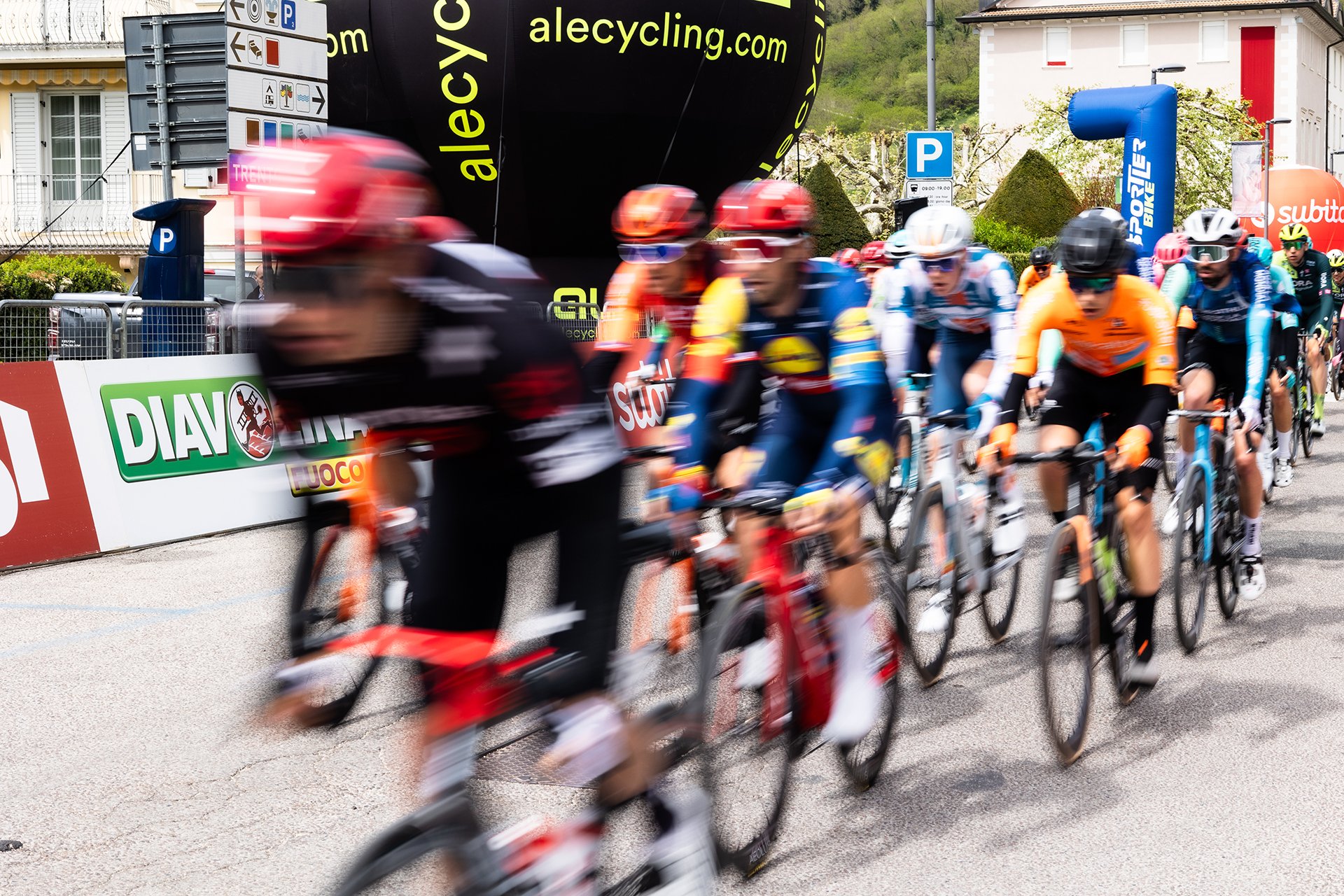 Cyclists competing in a race, blurred motion, riding on a street lined with banners and signs, with a crowd in the background.