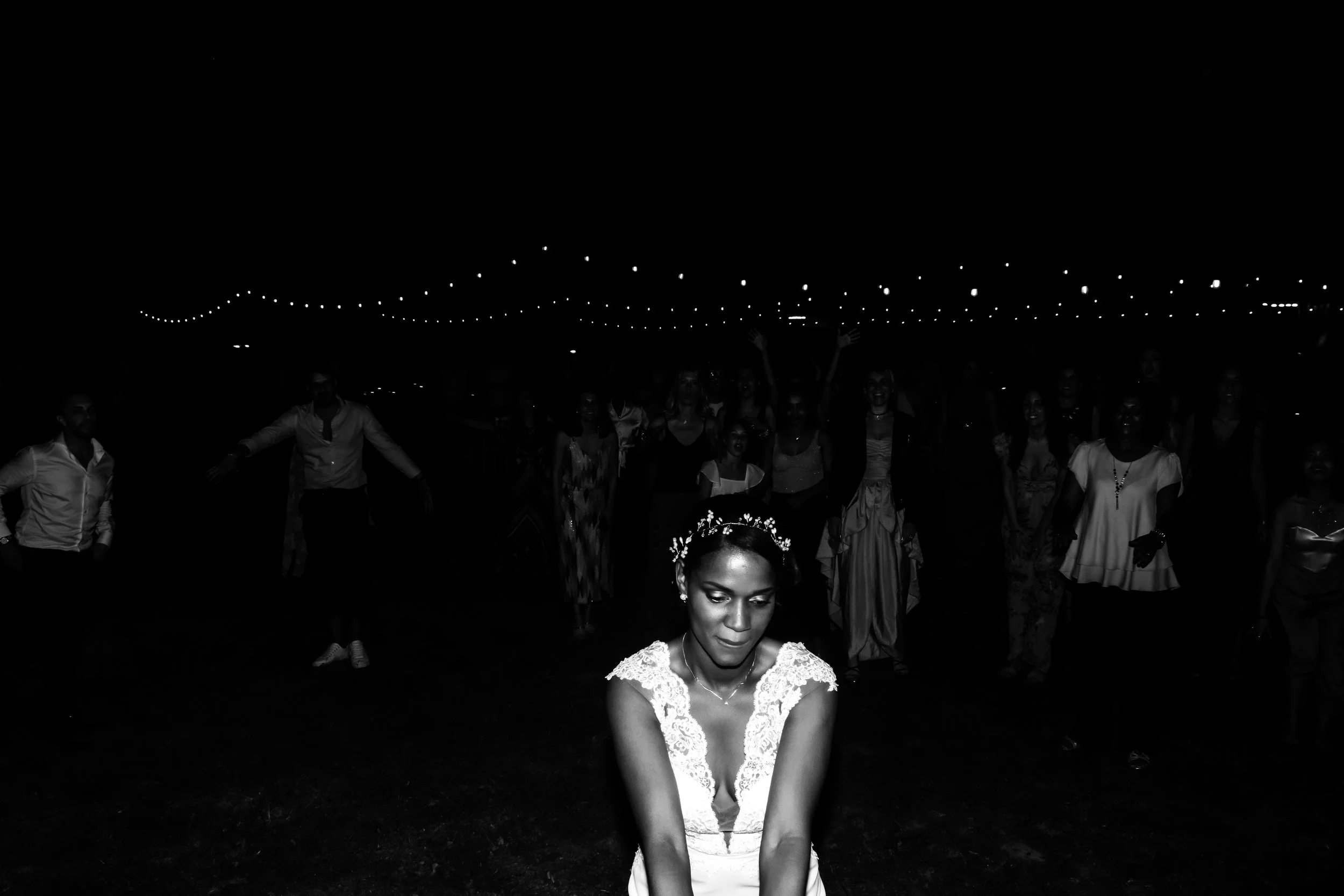 A woman in a white dress with intricate lace detail and a floral headpiece is in the foreground at a nighttime outdoor event. In the background, a group of people is gathered, with some raising their arms, and string lights are overhead, illuminating