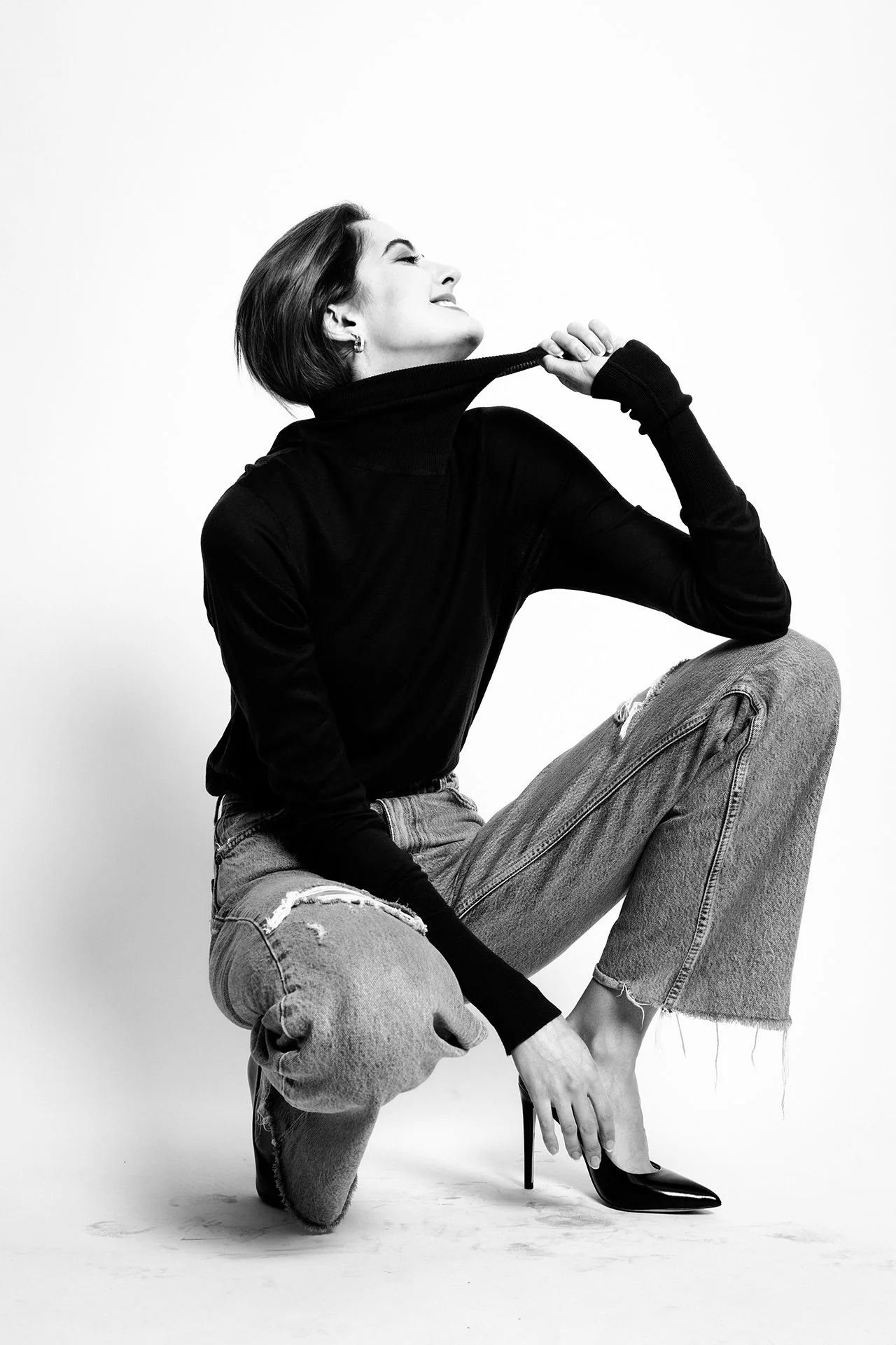 A woman with short hair wearing a black turtleneck, ripped jeans, and high heels, posing against a plain white background.