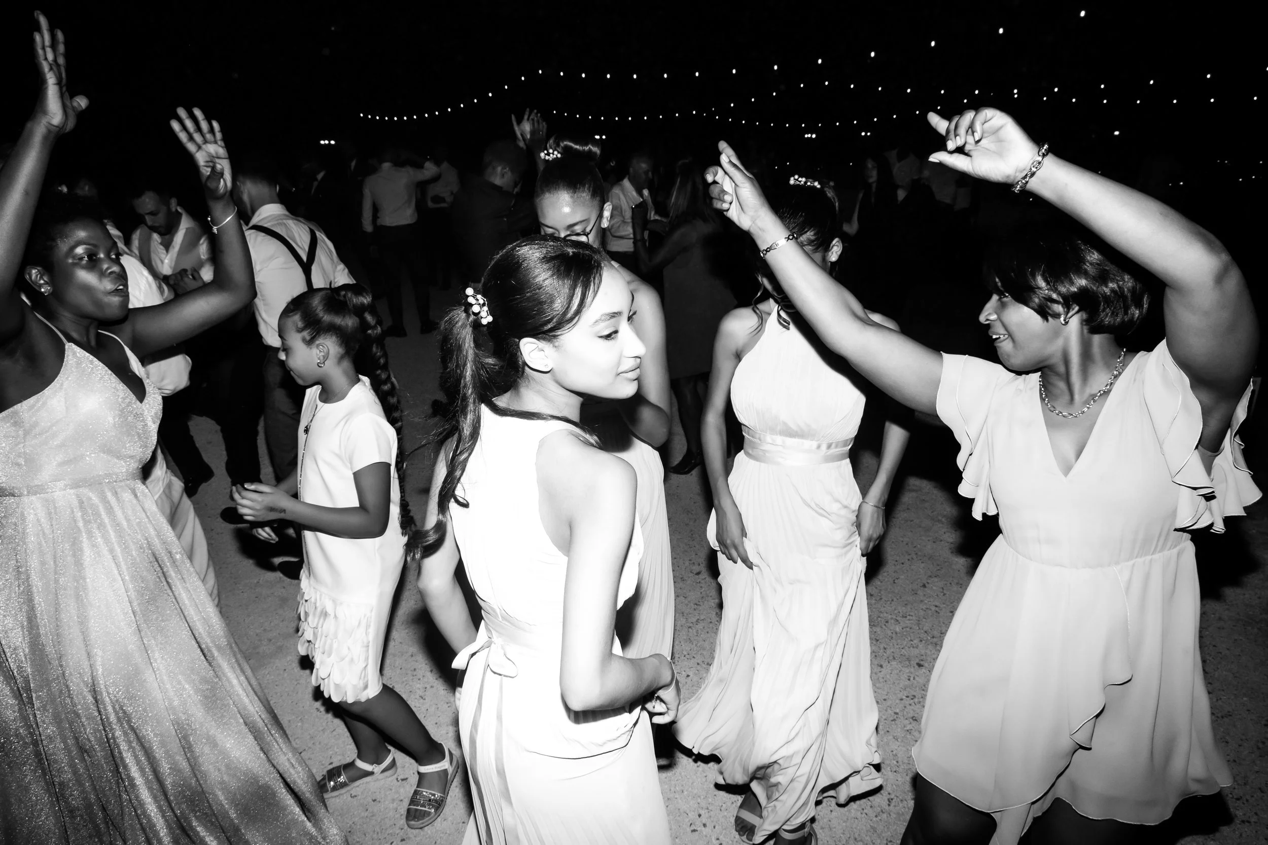Women and girls dancing at an indoor party or celebration, illuminated by string lights overhead, in black and white.
