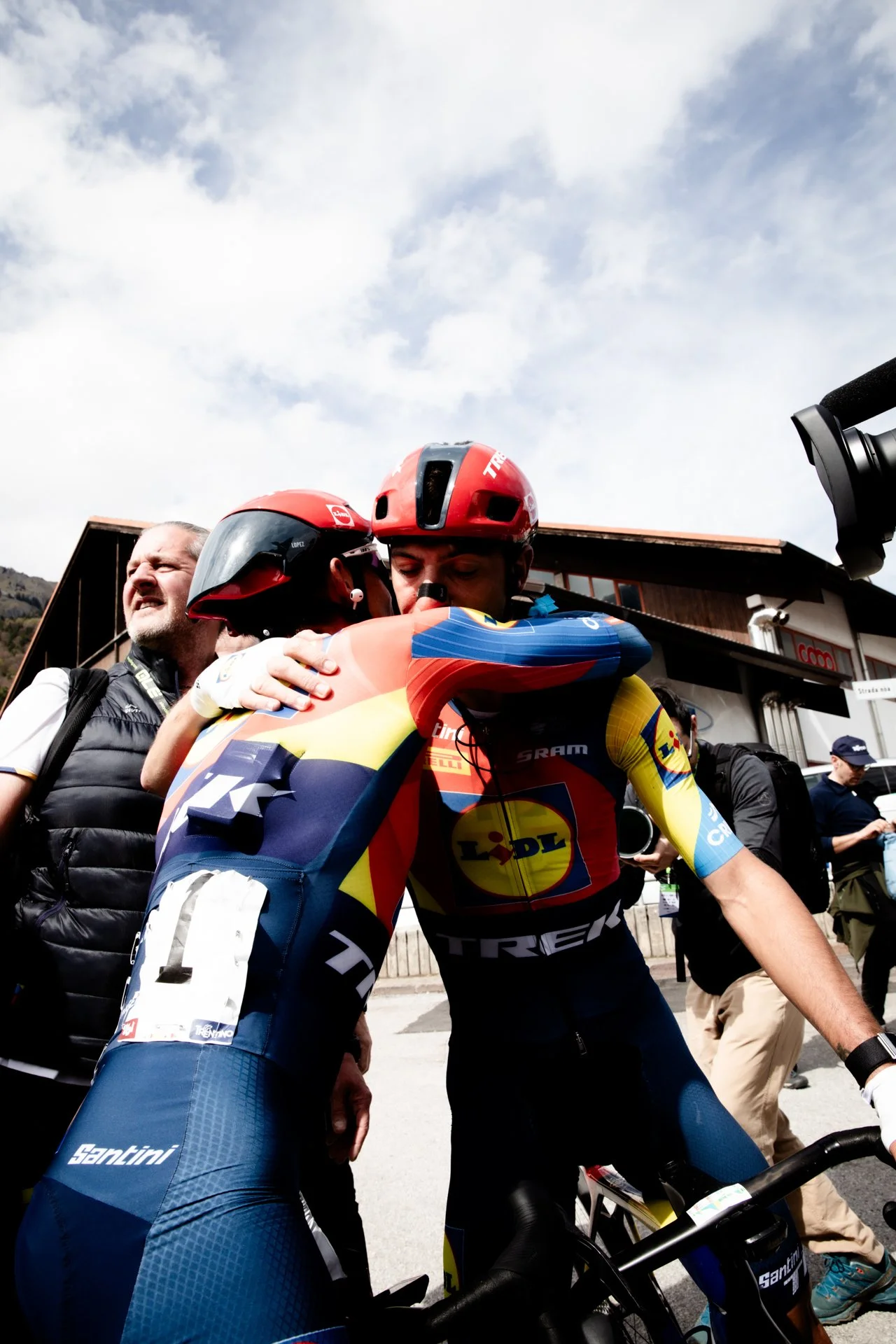 Cyclist in colorful team uniform hugging another cyclist with a red helmet, while other people are nearby, outdoors under a cloudy sky.