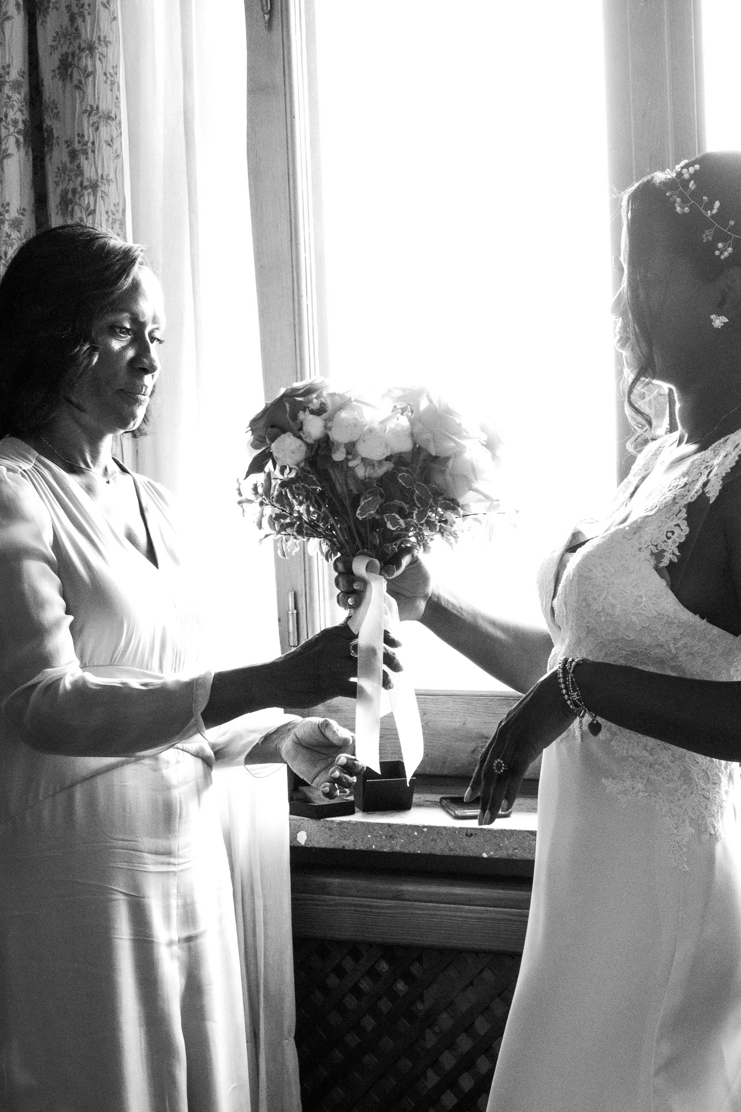 A black and white photo of two women exchanging a bouquet of flowers indoors, with a bright window in the background.