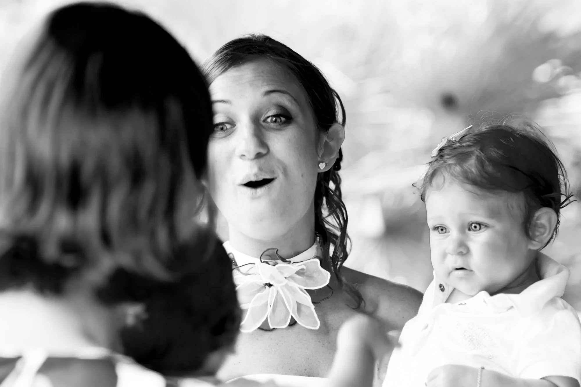 A woman holding a baby, engaging in conversation with another person, with her mouth open as if speaking, in an outdoor setting.
