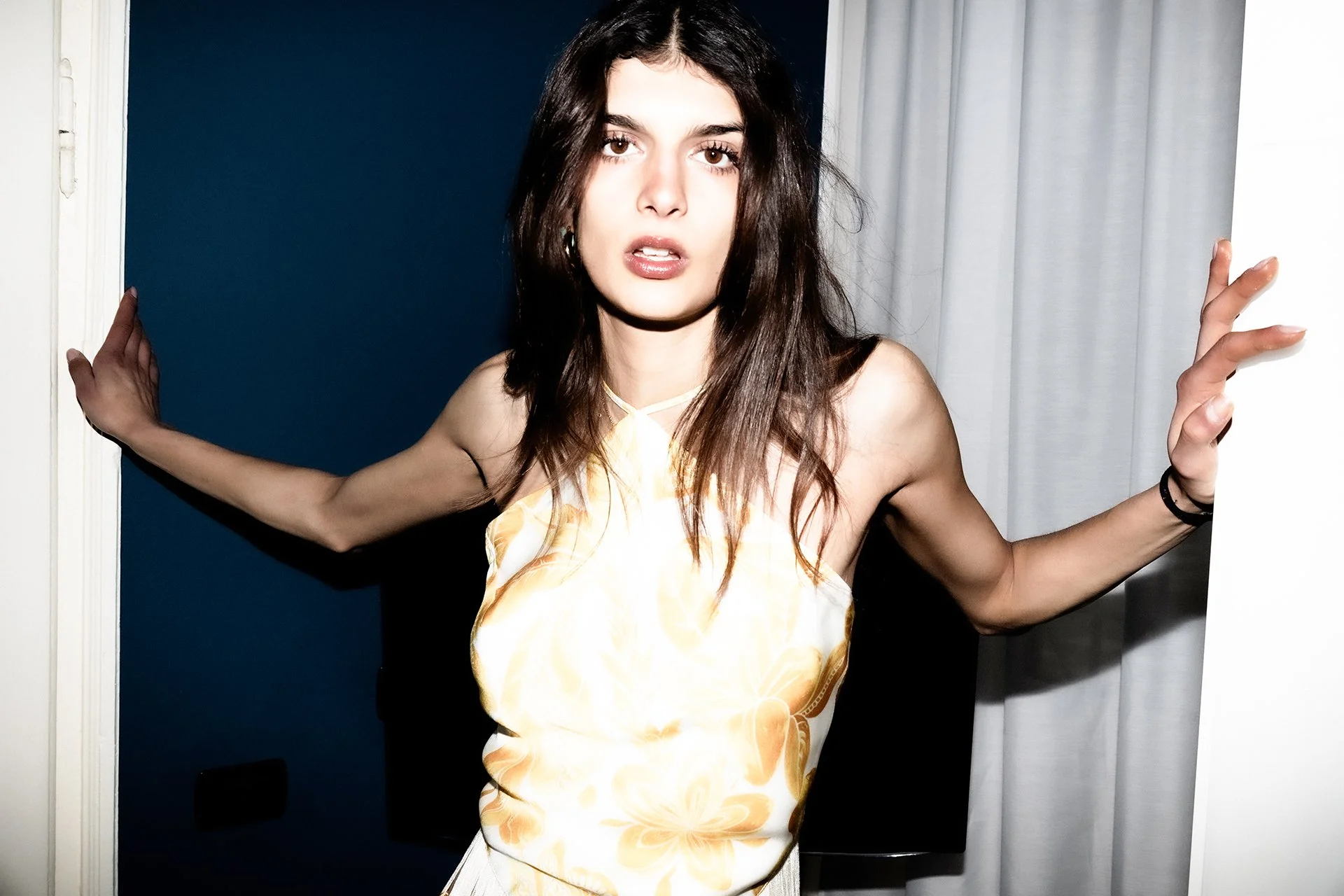 A woman with dark hair and light skin holding open a door with both hands, wearing a light-colored dress with an orange floral pattern, with white curtains in the background.